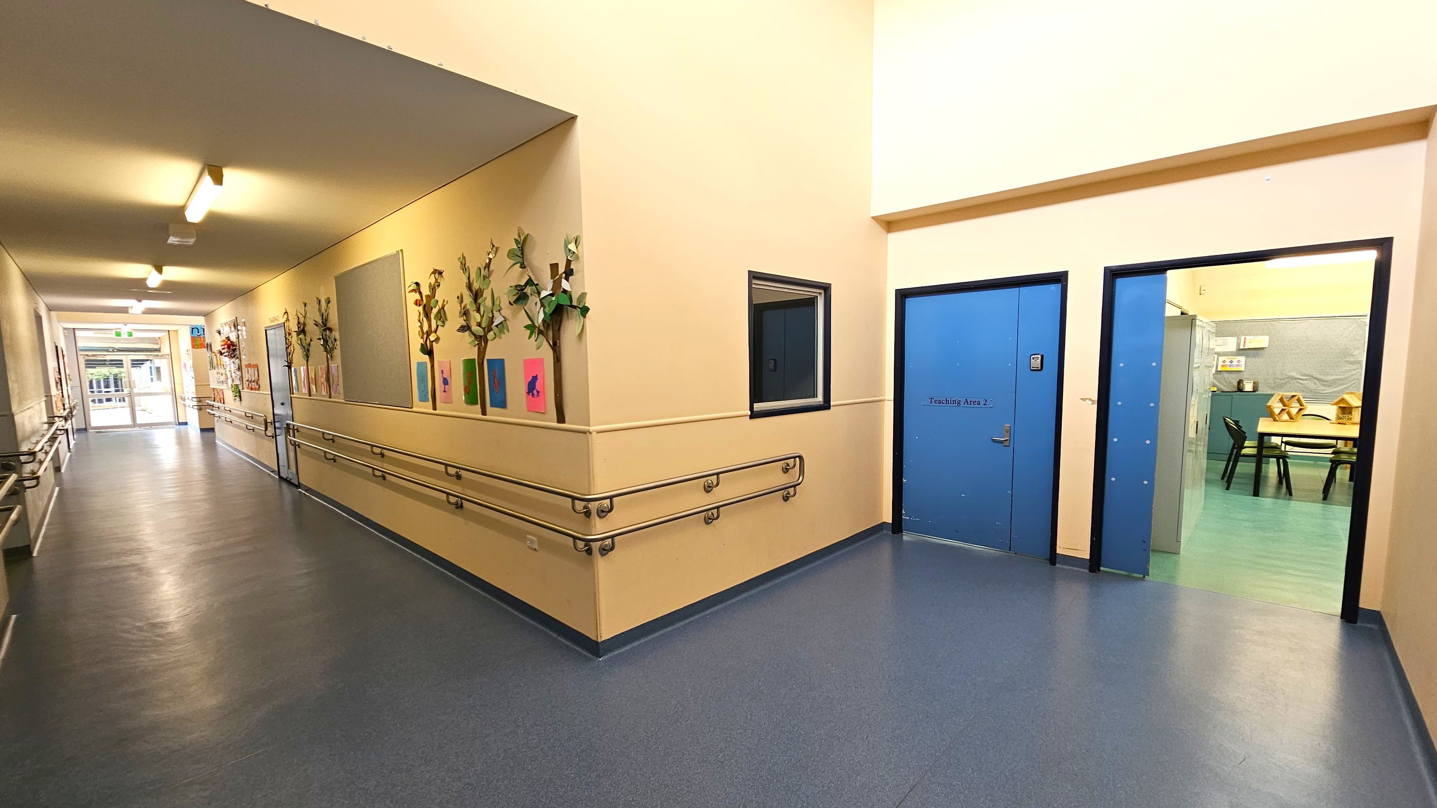 A corridor with a blue floor and cream walls. On the left, the wall is adorned with colourful artwork and decorative plants. Handrails run along both sides. At the end of the corridor, there is a glass door leading outside. On the right, there are two blue doors, one labelled "Teaching Area 2," and an open doorway revealing a room with tables and chairs. The ceiling is cream with fluorescent lights. A corridor with a blue floor and cream walls. On the left, the wall is adorned with colourful artwork and decorative plants. Handrails run along both sides. At the end of the corridor, there is a glass door leading outside. On the right, there are two blue doors, one labelled "Teaching Area 2," and an open doorway revealing a room with tables and chairs. The ceiling is cream with fluorescent lights.