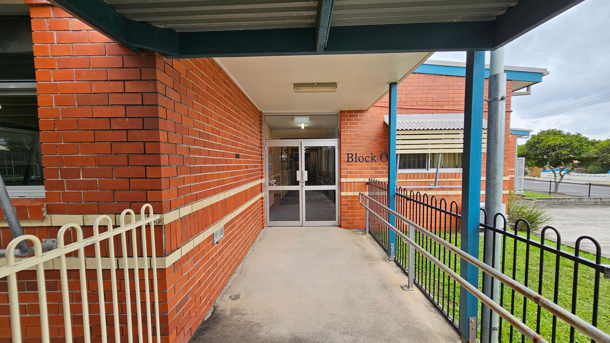 A brick building entrance with a sign reading "Block O" above the glass double doors. The left side features a red brick wall with a horizontal beige stripe and a window. On the right, there's a black metal fence and a grassy area. A blue metal post supports the overhanging roof. The concrete pathway leads to the entrance, and the ceiling is plain with a light fixture. In the background, there's a tree and a paved area. A brick building entrance with a sign reading "Block O" above the glass double doors. The left side features a red brick wall with a horizontal beige stripe and a window. On the right, there's a black metal fence and a grassy area. A blue metal post supports the overhanging roof. The concrete pathway leads to the entrance, and the ceiling is plain with a light fixture. In the background, there's a tree and a paved area.
