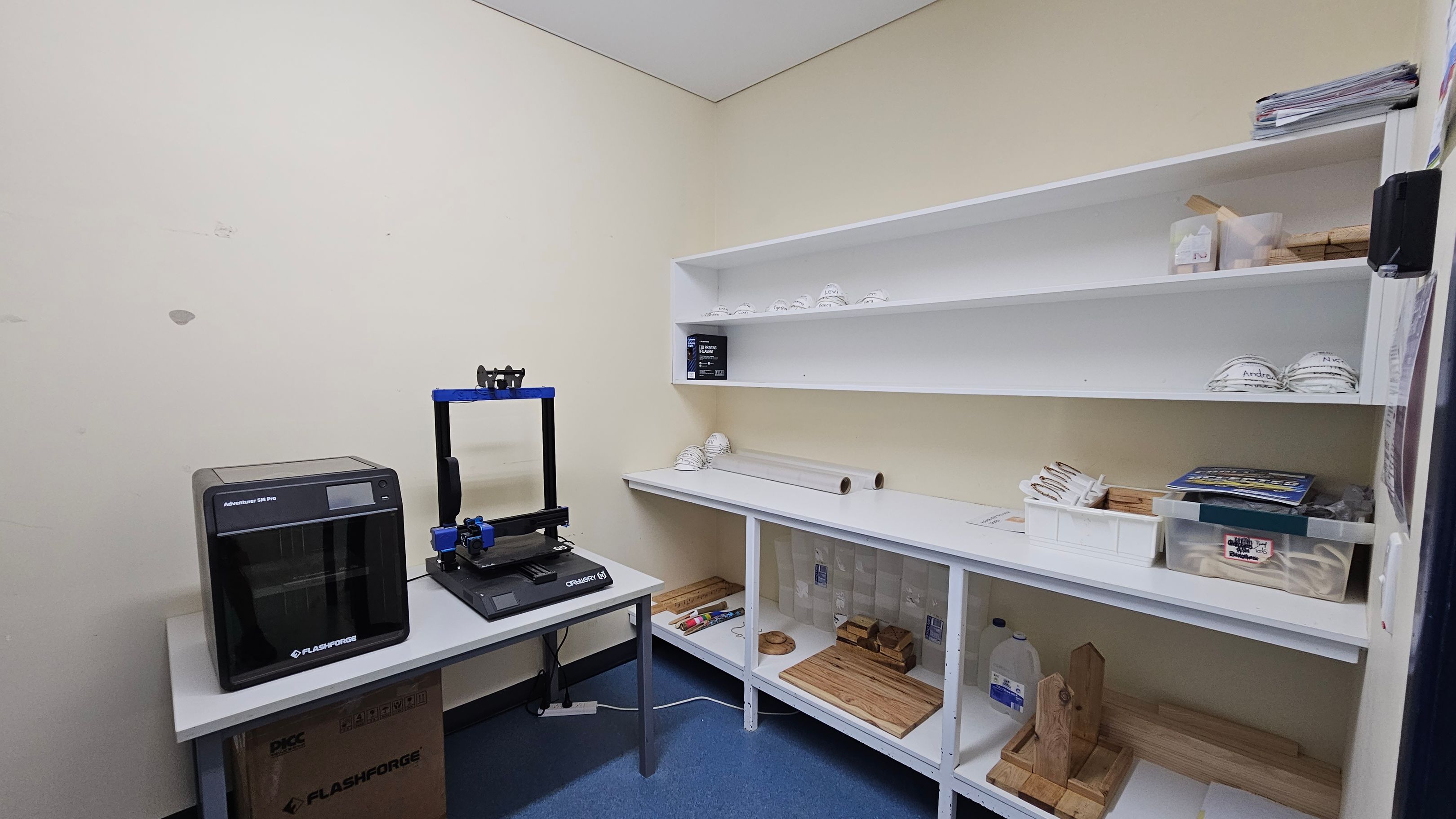 A small room with a blue floor and a white ceiling. On the left, there is a 3D printer on a table, with a cardboard box underneath. In the middle, white shelves hold various items, including stacked white objects, a box, and rolls of material. On the right, more shelves contain trays, books, and wooden items. The walls are a light colour, and the room appears organised for storage or work. A small room with a blue floor and a white ceiling. On the left, there is a 3D printer on a table, with a cardboard box underneath. In the middle, white shelves hold various items, including stacked white objects, a box, and rolls of material. On the right, more shelves contain trays, books, and wooden items. The walls are a light colour, and the room appears organised for storage or work.