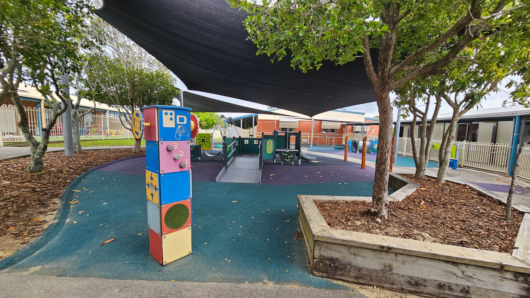 A playground with a colourful interactive panel on the left, featuring various shapes and numbers. In the centre, there's a play structure with slides and climbing elements. The ground is covered with a blue and green rubber surface. Trees with green foliage are on the right, and a wooden planter box is visible. Overhead, a large black shade cloth provides cover. In the background, there are buildings with brick and metal exteriors, and a white fence runs along the perimeter. A playground with a colourful interactive panel on the left, featuring various shapes and numbers. In the centre, there's a play structure with slides and climbing elements. The ground is covered with a blue and green rubber surface. Trees with green foliage are on the right, and a wooden planter box is visible. Overhead, a large black shade cloth provides cover. In the background, there are buildings with brick and metal exteriors, and a white fence runs along the perimeter.
