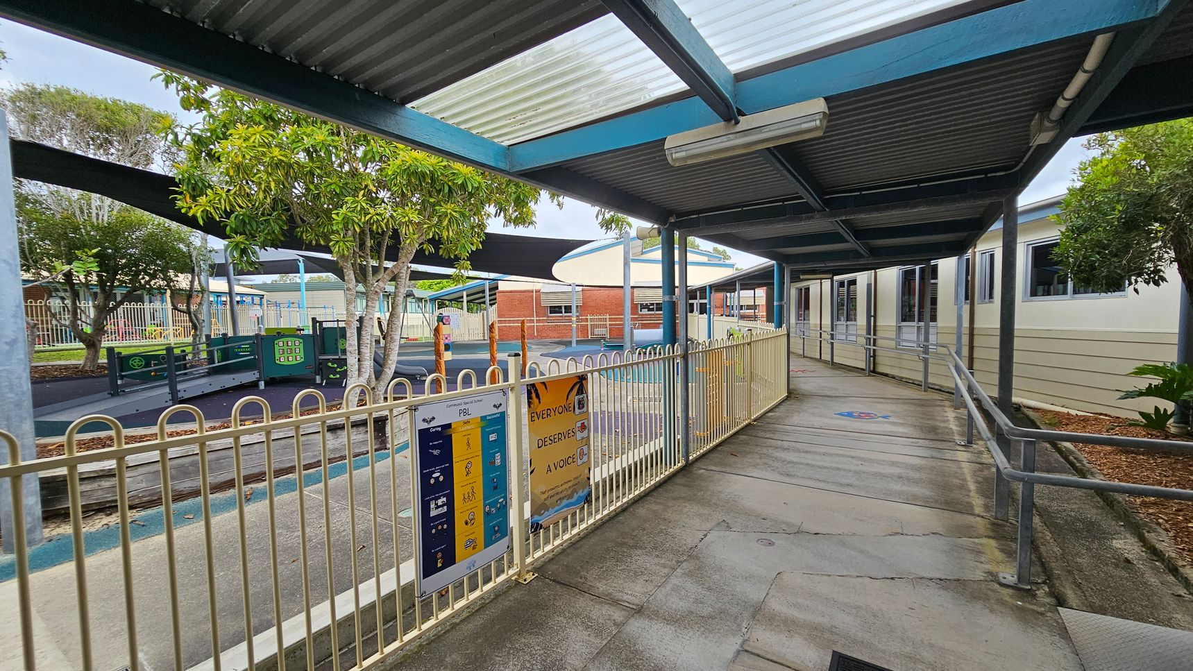 A covered walkway with a corrugated metal roof and transparent panels runs alongside a building with multiple windows on the right. A metal railing lines the path, with signs attached. On the left, a fenced playground area features trees, play equipment, and shade structures. The ground is a mix of concrete and rubberised surfaces. In the background, brick buildings are visible, and the area is surrounded by more fencing and greenery. A covered walkway with a corrugated metal roof and transparent panels runs alongside a building with multiple windows on the right. A metal railing lines the path, with signs attached. On the left, a fenced playground area features trees, play equipment, and shade structures. The ground is a mix of concrete and rubberised surfaces. In the background, brick buildings are visible, and the area is surrounded by more fencing and greenery.