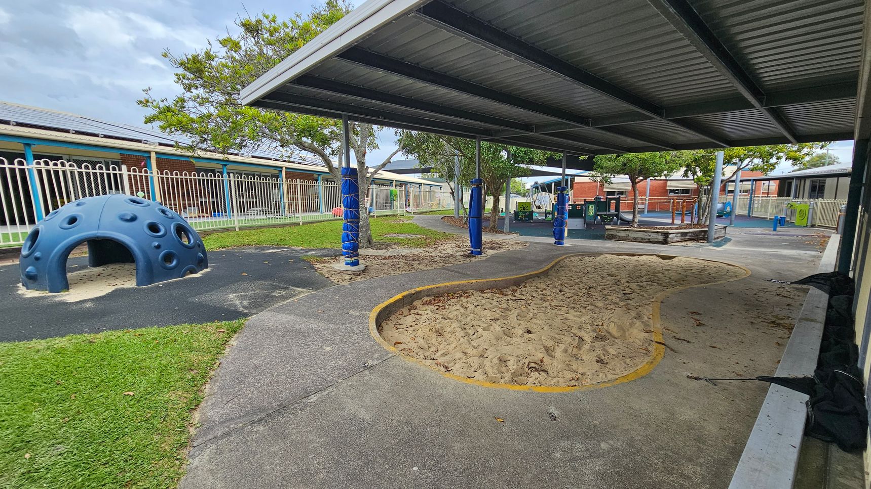A playground with a blue dome-shaped climbing structure on the left, surrounded by grass and a paved area. In the centre, there's a sandpit with a yellow border. The right side features a covered area with a metal roof supported by poles wrapped in blue padding. In the background, there are trees, a fence, and school buildings with blue and red accents. The ground is a mix of grass, sand, and pavement. A playground with a blue dome-shaped climbing structure on the left, surrounded by grass and a paved area. In the centre, there's a sandpit with a yellow border. The right side features a covered area with a metal roof supported by poles wrapped in blue padding. In the background, there are trees, a fence, and school buildings with blue and red accents. The ground is a mix of grass, sand, and pavement.