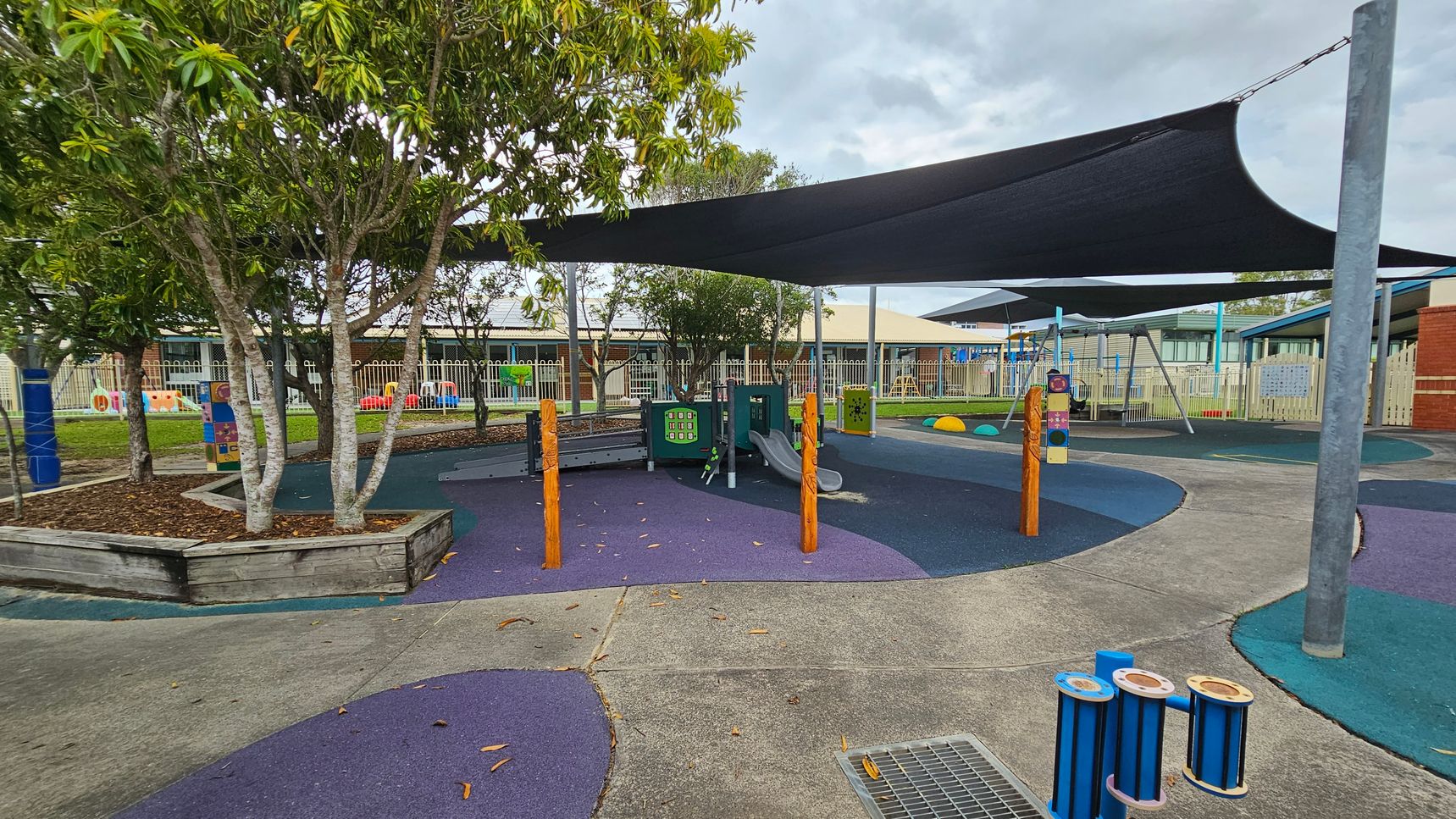 A playground with a variety of equipment, including a slide and interactive panels, is covered by large black shade sails. The ground is a mix of purple and blue soft surfaces. On the left, there are trees in a raised wooden planter. In the background, a fence encloses the area, with a building visible beyond it. The foreground features a concrete path and a small blue musical instrument. A playground with a variety of equipment, including a slide and interactive panels, is covered by large black shade sails. The ground is a mix of purple and blue soft surfaces. On the left, there are trees in a raised wooden planter. In the background, a fence encloses the area, with a building visible beyond it. The foreground features a concrete path and a small blue musical instrument.