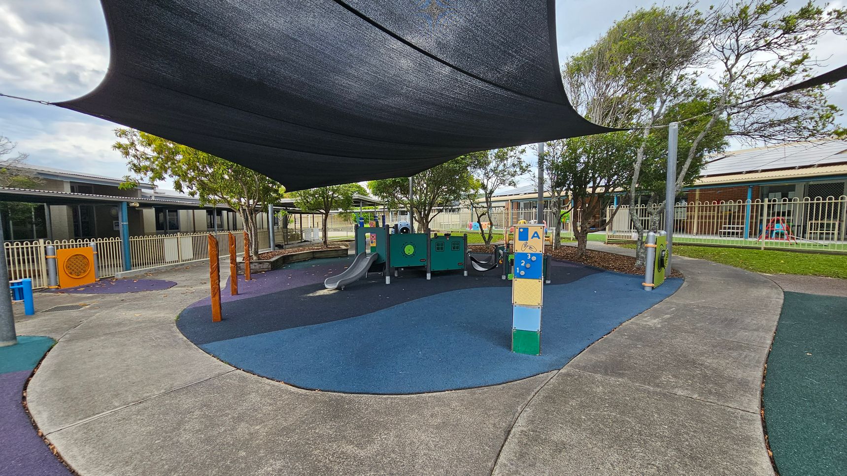 A playground with a blue and green rubber surface. In the centre, there is a play structure with slides and climbing features. A large black shade sail is overhead. Trees and a fence line the background, with a building visible behind them. The ground is a mix of blue and green pathways, and there are wooden posts and interactive play panels scattered around. A playground with a blue and green rubber surface. In the centre, there is a play structure with slides and climbing features. A large black shade sail is overhead. Trees and a fence line the background, with a building visible behind them. The ground is a mix of blue and green pathways, and there are wooden posts and interactive play panels scattered around.