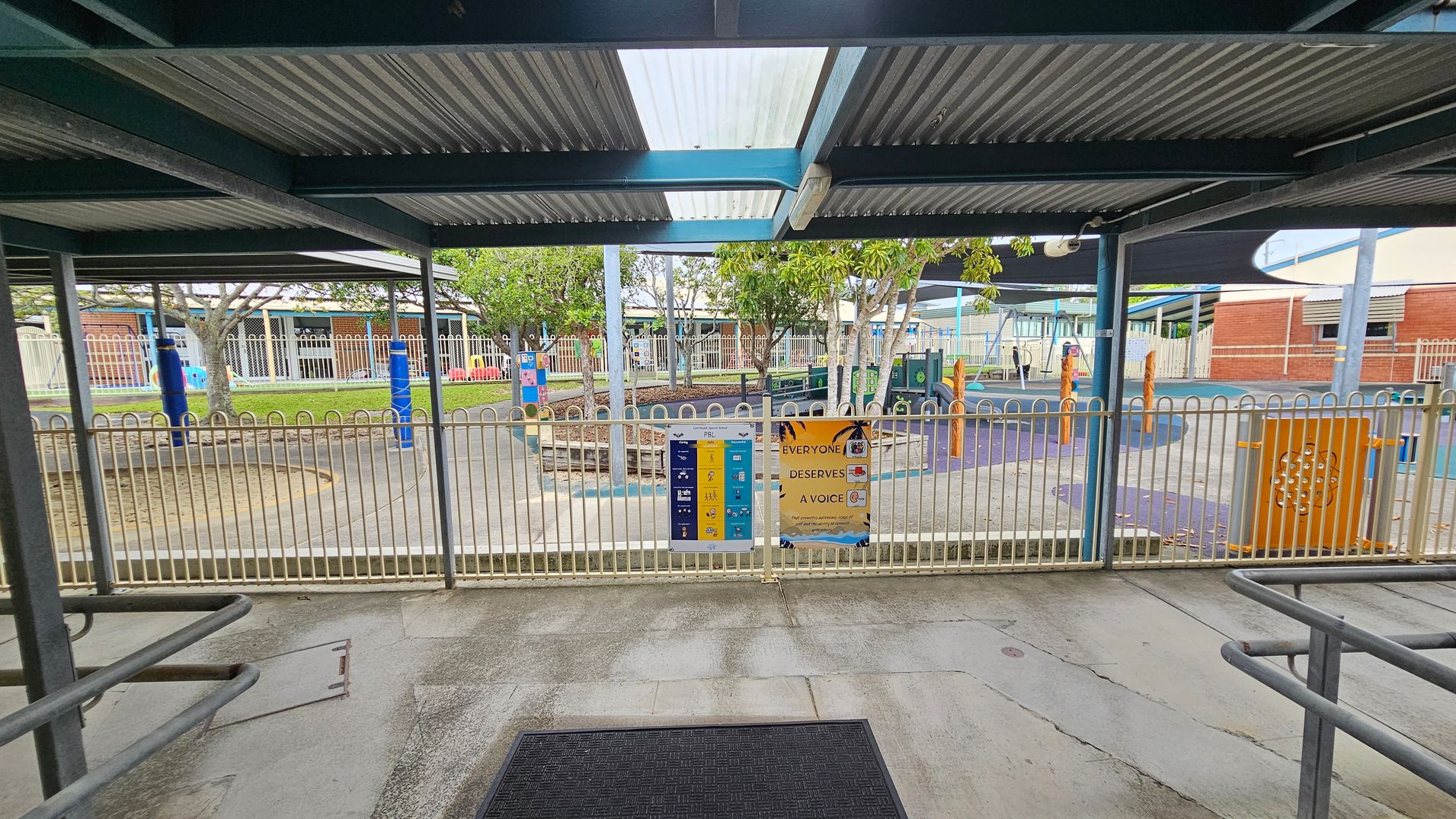 A covered outdoor area with a corrugated metal roof and a black mat on the concrete floor. Beyond the metal fence, there is a playground with colourful equipment, including blue and orange structures. Trees provide some shade, and there are signs on the fence with text and graphics. In the background, there are buildings with brick and metal elements, and a shaded area with a dark canopy. A covered outdoor area with a corrugated metal roof and a black mat on the concrete floor. Beyond the metal fence, there is a playground with colourful equipment, including blue and orange structures. Trees provide some shade, and there are signs on the fence with text and graphics. In the background, there are buildings with brick and metal elements, and a shaded area with a dark canopy.