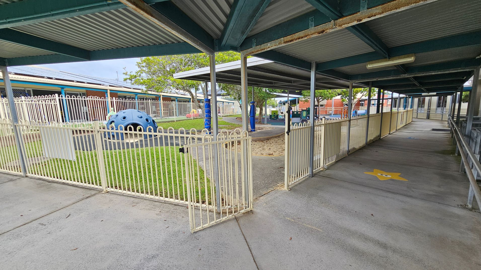 A covered walkway with a corrugated metal roof and metal support beams. On the left, there's a fenced playground area with a blue climbing structure on grass. The fence is white with a gate slightly open. In the background, trees and a brick building are visible. On the right, the walkway has a yellow star painted on the concrete floor and is bordered by a railing. The ceiling has a fluorescent light fixture. A covered walkway with a corrugated metal roof and metal support beams. On the left, there's a fenced playground area with a blue climbing structure on grass. The fence is white with a gate slightly open. In the background, trees and a brick building are visible. On the right, the walkway has a yellow star painted on the concrete floor and is bordered by a railing. The ceiling has a fluorescent light fixture.