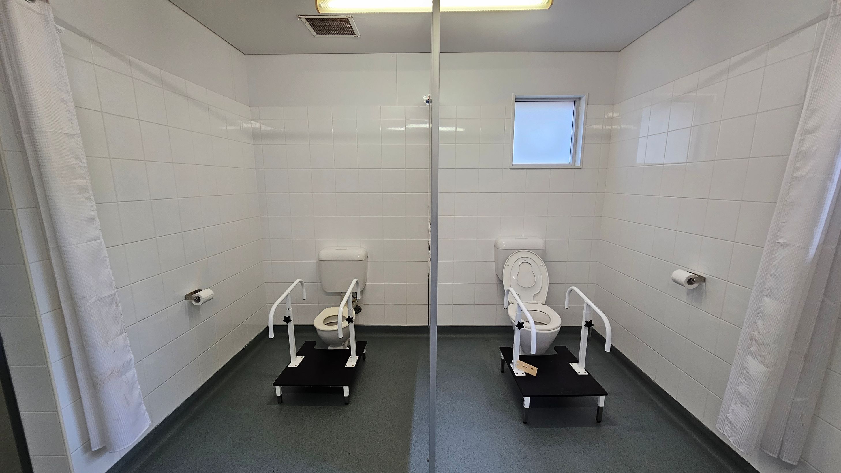 Two toilets are positioned side by side, separated by a glass partition. Each toilet is equipped with support rails and a raised platform. The walls are covered with white tiles, and there is a small window on the right side. Toilet paper holders are mounted on the walls next to each toilet. The floor is grey, and the ceiling is white with a light fixture and vent. A white curtain is partially visible on the left. Two toilets are positioned side by side, separated by a glass partition. Each toilet is equipped with support rails and a raised platform. The walls are covered with white tiles, and there is a small window on the right side. Toilet paper holders are mounted on the walls next to each toilet. The floor is grey, and the ceiling is white with a light fixture and vent. A white curtain is partially visible on the left.