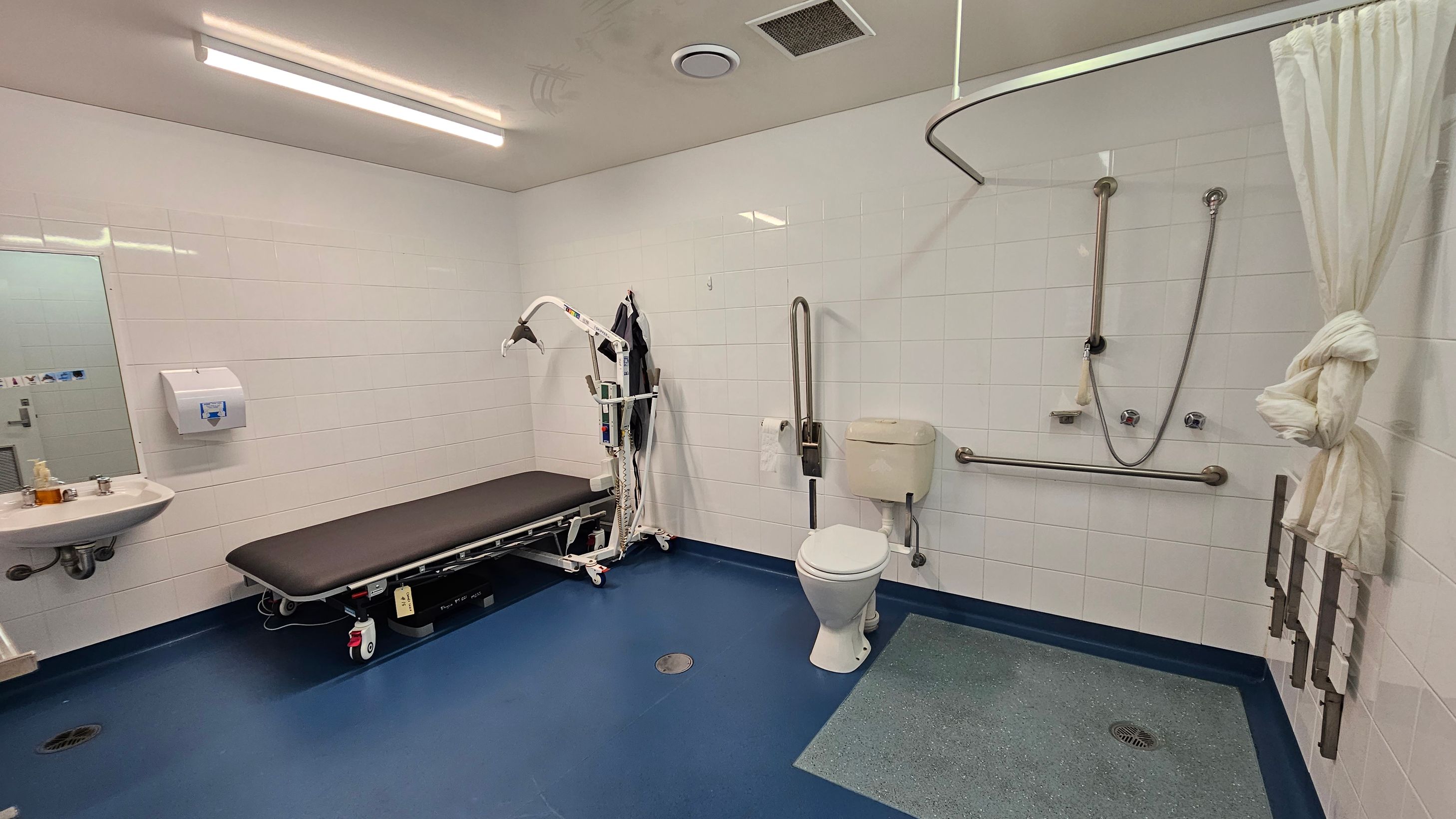 A spacious, accessible bathroom with white tiled walls and a blue floor. On the left, there's a sink with a mirror above it and a paper towel dispenser. A black cushioned bench with a hoist is positioned nearby. On the right, a toilet with grab bars is adjacent to a shower area with a curtain tied to the side. The ceiling has fluorescent lighting and ventilation fixtures. A spacious, accessible bathroom with white tiled walls and a blue floor. On the left, there's a sink with a mirror above it and a paper towel dispenser. A black cushioned bench with a hoist is positioned nearby. On the right, a toilet with grab bars is adjacent to a shower area with a curtain tied to the side. The ceiling has fluorescent lighting and ventilation fixtures.