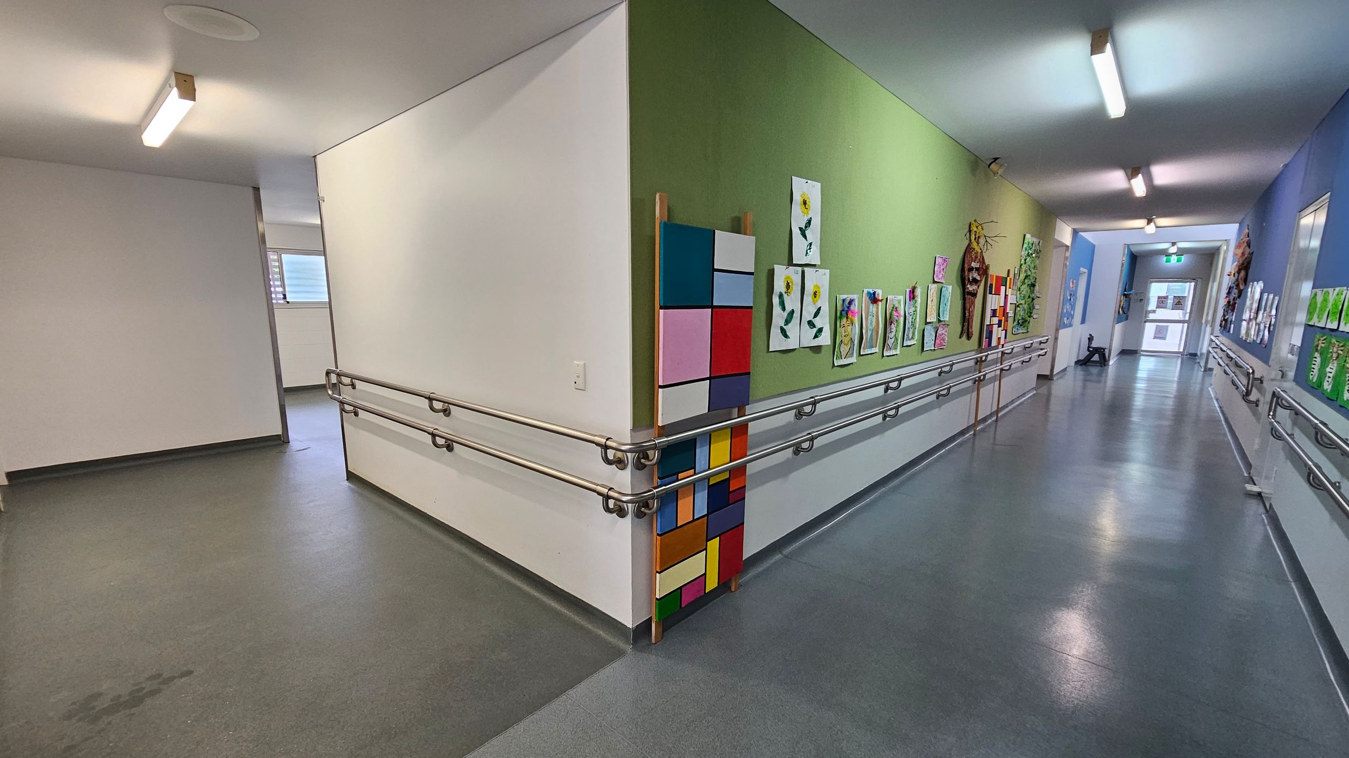 A corridor with a green and blue wall on the right, displaying various colourful artworks, including paintings of flowers and abstract designs. The left side has a plain white wall with a window in the background. The floor is grey, and there are metal handrails along the walls. The ceiling features fluorescent lights. At the end of the corridor, there is a door with a glass panel. A corridor with a green and blue wall on the right, displaying various colourful artworks, including paintings of flowers and abstract designs. The left side has a plain white wall with a window in the background. The floor is grey, and there are metal handrails along the walls. The ceiling features fluorescent lights. At the end of the corridor, there is a door with a glass panel.