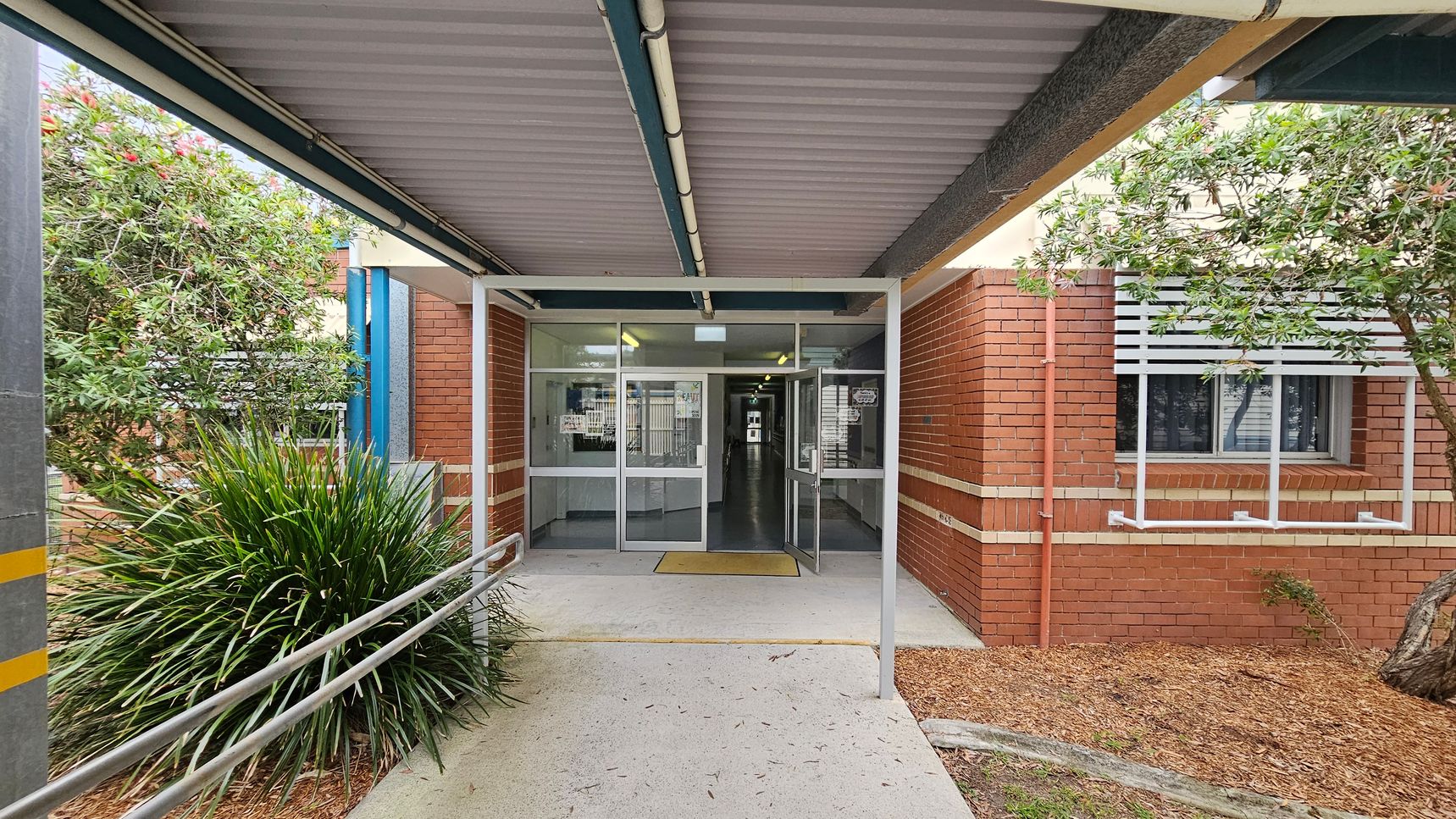 A covered walkway leads to a building entrance with glass doors. The structure has a corrugated metal roof and a metal railing on the left. On the left, there is a bushy plant and a tree, while on the right, a brick wall with a window featuring white slats is visible. The ground is a mix of concrete and mulch. Inside, fluorescent lights illuminate a hallway. A covered walkway leads to a building entrance with glass doors. The structure has a corrugated metal roof and a metal railing on the left. On the left, there is a bushy plant and a tree, while on the right, a brick wall with a window featuring white slats is visible. The ground is a mix of concrete and mulch. Inside, fluorescent lights illuminate a hallway.