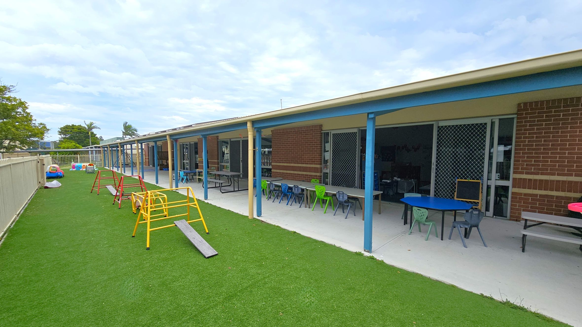A playground with artificial grass on the left, featuring colourful play equipment like small climbing frames and toy cars. On the right, a building with a covered patio area has tables and chairs in various colours. The building has brick walls and sliding doors with mesh screens. A fence runs along the left side, and there are trees in the background. A playground with artificial grass on the left, featuring colourful play equipment like small climbing frames and toy cars. On the right, a building with a covered patio area has tables and chairs in various colours. The building has brick walls and sliding doors with mesh screens. A fence runs along the left side, and there are trees in the background.