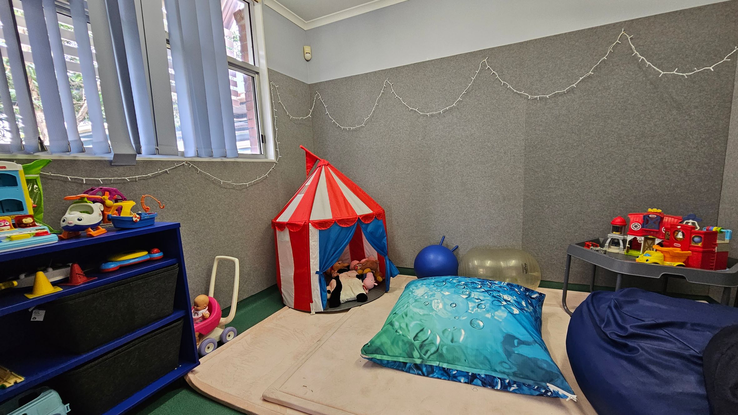 A children's playroom with a green carpeted floor. On the left, a blue shelf holds various toys, including a toy helicopter and a doll in a pink stroller. In the centre, a red and white striped play tent contains stuffed animals. A large blue cushion and a dark blue bean bag are on the floor. On the right, a grey table holds more toys, including a red toy train. The walls are grey with string lights hanging, and there are vertical blinds on the window. A children's playroom with a green carpeted floor. On the left, a blue shelf holds various toys, including a toy helicopter and a doll in a pink stroller. In the centre, a red and white striped play tent contains stuffed animals. A large blue cushion and a dark blue bean bag are on the floor. On the right, a grey table holds more toys, including a red toy train. The walls are grey with string lights hanging, and there are vertical blinds on the window.