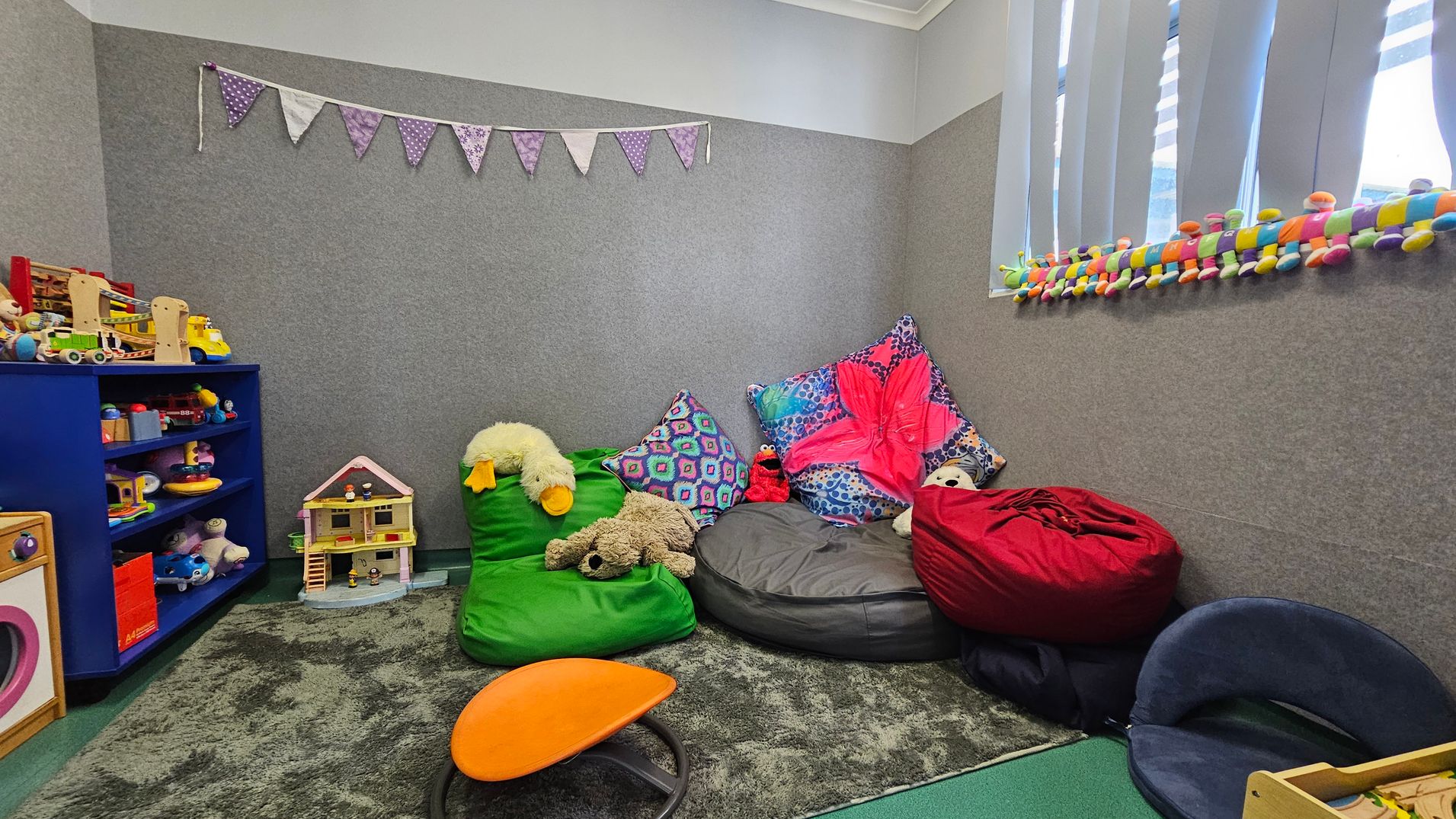 A cosy playroom with grey walls and a green carpet. On the left, a blue shelf holds various toys, including a dollhouse and colourful play items. In the middle, bean bags in green, grey, and red are adorned with plush toys and vibrant cushions. On the right, a window with vertical blinds has a colourful caterpillar toy on the sill. A string of purple bunting hangs on the wall, adding a playful touch. An orange stool is on the floor. A cosy playroom with grey walls and a green carpet. On the left, a blue shelf holds various toys, including a dollhouse and colourful play items. In the middle, bean bags in green, grey, and red are adorned with plush toys and vibrant cushions. On the right, a window with vertical blinds has a colourful caterpillar toy on the sill. A string of purple bunting hangs on the wall, adding a playful touch. An orange stool is on the floor.