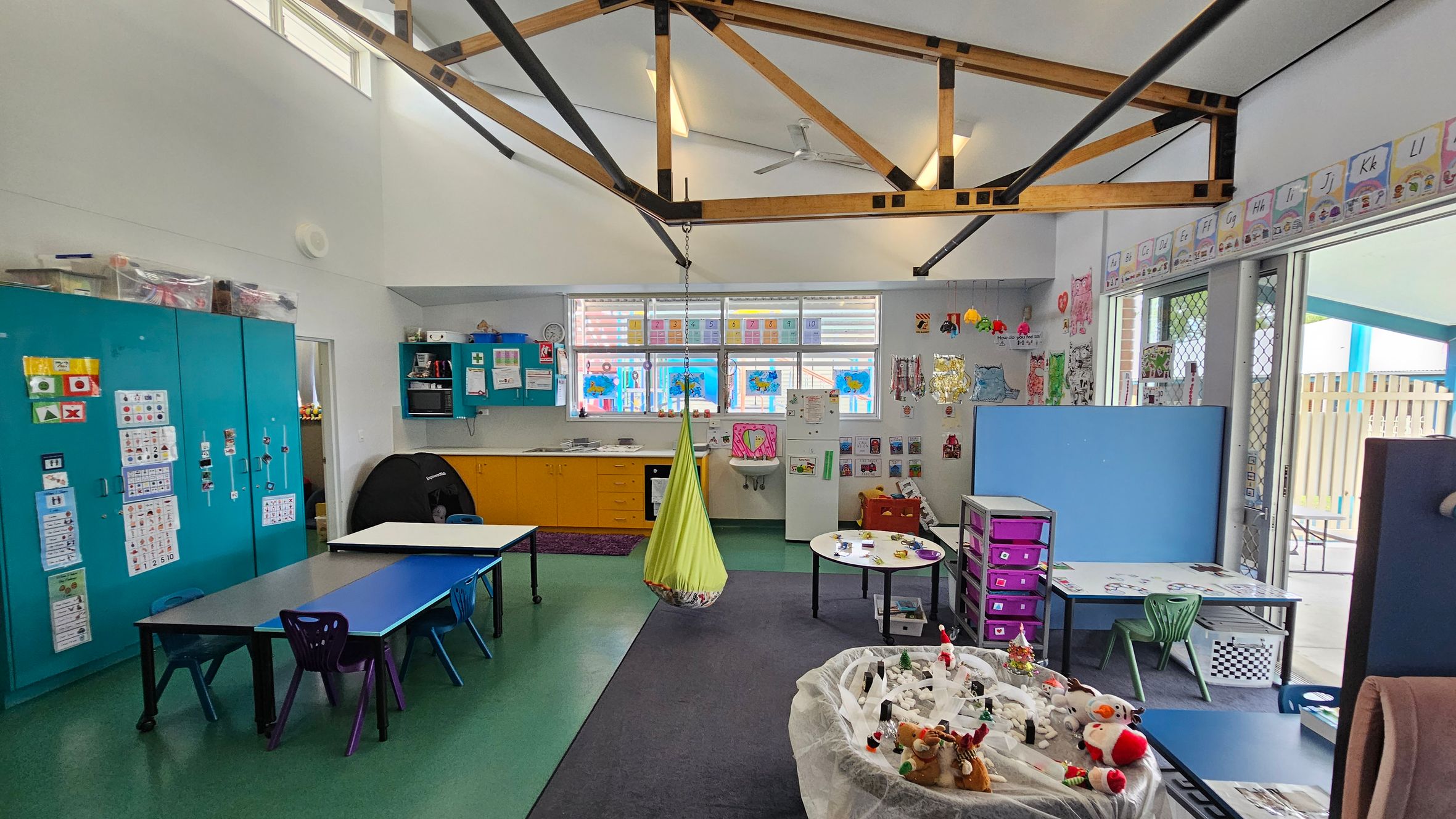 A classroom with a high ceiling featuring exposed wooden beams. On the left, there are blue cabinets with educational posters. In the centre, a green hanging chair is suspended from the ceiling. The back wall has windows with colourful artwork and yellow cabinets below. On the right, tables with chairs and a play area with toys are visible. The floor is a mix of green and grey, and the walls are decorated with various educational materials. A classroom with a high ceiling featuring exposed wooden beams. On the left, there are blue cabinets with educational posters. In the centre, a green hanging chair is suspended from the ceiling. The back wall has windows with colourful artwork and yellow cabinets below. On the right, tables with chairs and a play area with toys are visible. The floor is a mix of green and grey, and the walls are decorated with various educational materials.