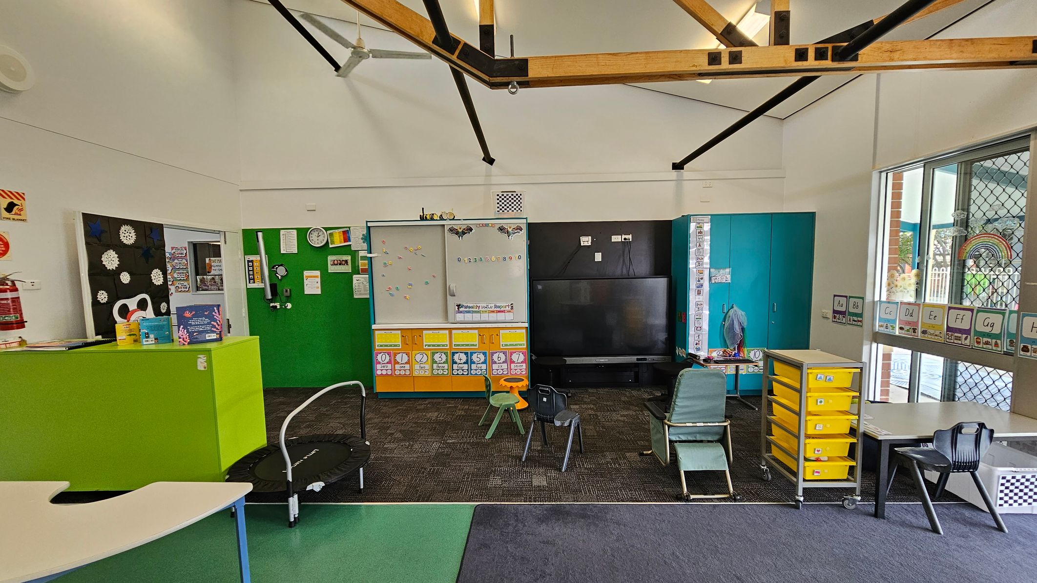 Classroom with a high ceiling featuring exposed wooden beams. The left side has a green partition with educational materials and a fire extinguisher. In the centre, a whiteboard with colourful magnets and a large black TV. Chairs and a small trampoline are on the carpeted floor. The right side has a blue cabinet, a table with yellow drawers, and a window with alphabet cards. The floor is a mix of green and dark carpet. Classroom with a high ceiling featuring exposed wooden beams. The left side has a green partition with educational materials and a fire extinguisher. In the centre, a whiteboard with colourful magnets and a large black TV. Chairs and a small trampoline are on the carpeted floor. The right side has a blue cabinet, a table with yellow drawers, and a window with alphabet cards. The floor is a mix of green and dark carpet.