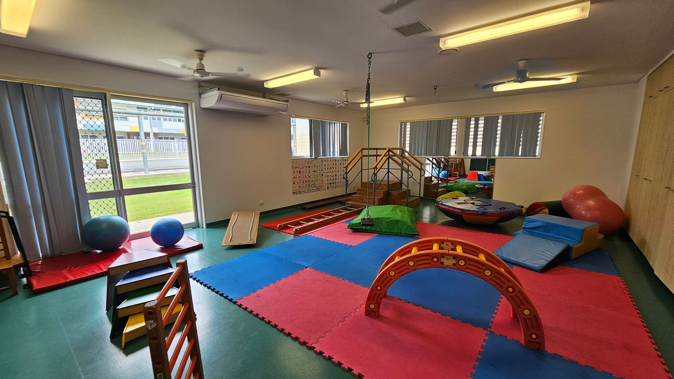 A children's playroom with a green floor and a white ceiling. On the left, there are large windows with vertical blinds, a mesh door, and two blue exercise balls on red mats. In the centre, colourful foam mats cover the floor, with a small wooden climbing structure and a red arch. On the right, there's a large red exercise ball, blue foam blocks, and a circular play platform. The back wall has a small staircase with a railing and a chain hanging from the ceiling. A children's playroom with a green floor and a white ceiling. On the left, there are large windows with vertical blinds, a mesh door, and two blue exercise balls on red mats. In the centre, colourful foam mats cover the floor, with a small wooden climbing structure and a red arch. On the right, there's a large red exercise ball, blue foam blocks, and a circular play platform. The back wall has a small staircase with a railing and a chain hanging from the ceiling.