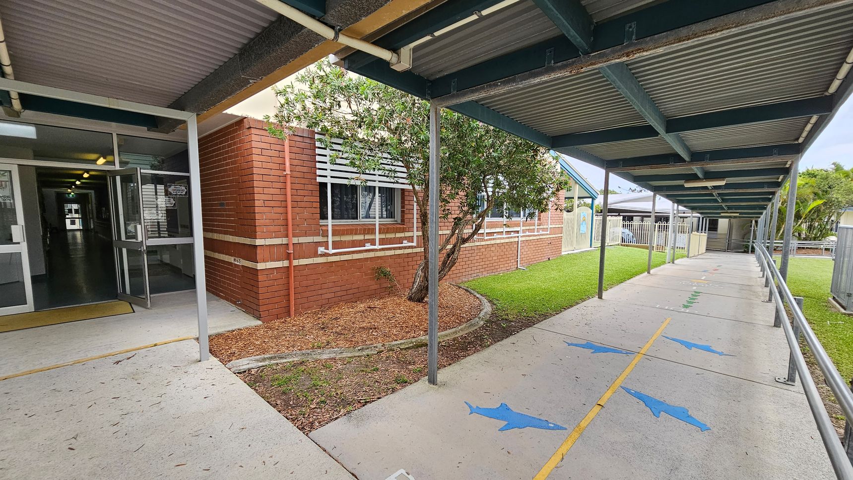 A covered walkway with a corrugated metal roof extends to the right, featuring a concrete path marked with blue dolphin shapes and a yellow line. On the left, a brick building with white window shutters and a small tree in a mulched area is visible. The entrance to the building has glass doors, leading to a lit interior hallway. The grassy area runs alongside the path, bordered by a fence in the background. A covered walkway with a corrugated metal roof extends to the right, featuring a concrete path marked with blue dolphin shapes and a yellow line. On the left, a brick building with white window shutters and a small tree in a mulched area is visible. The entrance to the building has glass doors, leading to a lit interior hallway. The grassy area runs alongside the path, bordered by a fence in the background.