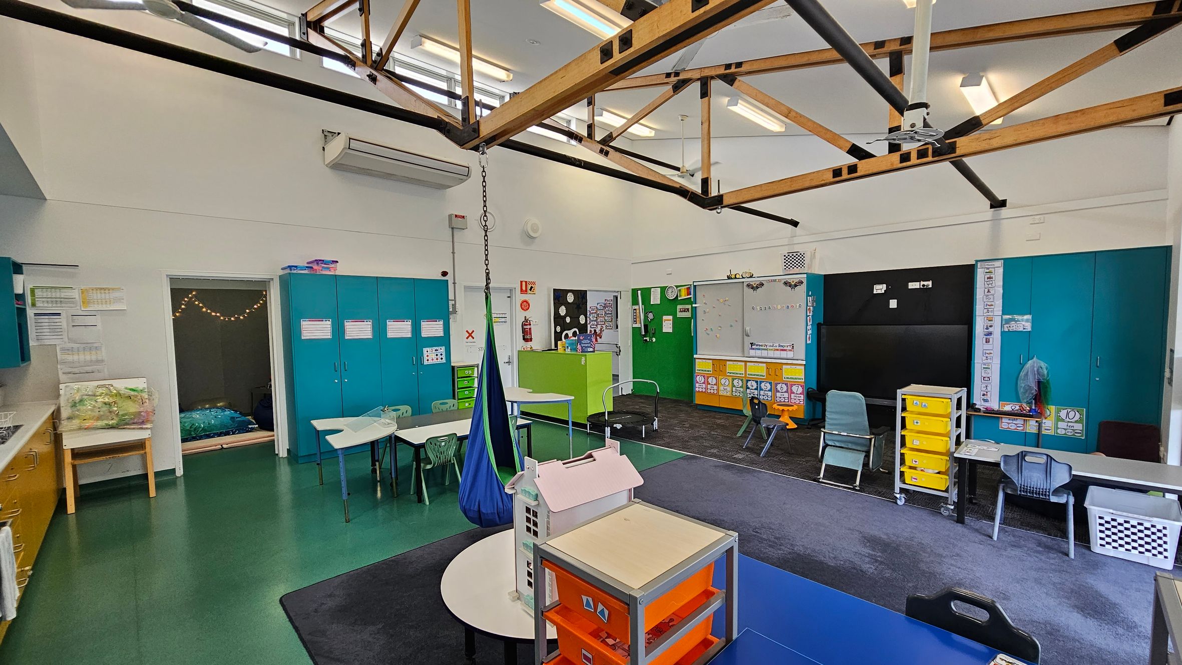 Classroom with green flooring and a high ceiling featuring exposed wooden beams. On the left, there are blue cabinets and a small room with string lights. In the centre, tables and chairs are arranged, with a blue hanging chair. The right side has a blackboard, colourful storage units, and more chairs. A small playhouse and a trolley with yellow drawers are in the foreground. The walls are decorated with educational materials. Classroom with green flooring and a high ceiling featuring exposed wooden beams. On the left, there are blue cabinets and a small room with string lights. In the centre, tables and chairs are arranged, with a blue hanging chair. The right side has a blackboard, colourful storage units, and more chairs. A small playhouse and a trolley with yellow drawers are in the foreground. The walls are decorated with educational materials.