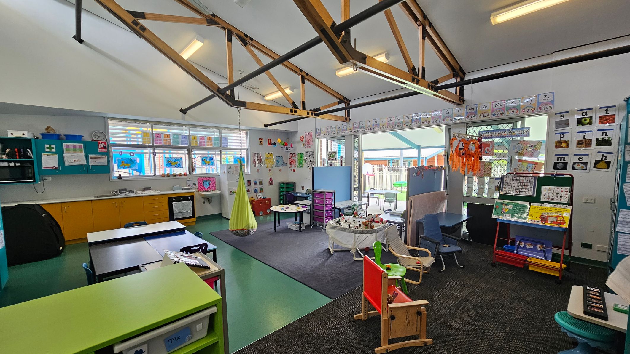 Classroom with a high ceiling featuring exposed beams and fluorescent lights. On the left, there are yellow cabinets and a sink beneath windows decorated with colourful artwork. In the centre, a small round table and a hanging green chair are on a carpeted area. The right side has a display board with educational materials and a variety of chairs. The walls are adorned with alphabet cards and children's artwork. The floor is a mix of green and dark carpet. Classroom with a high ceiling featuring exposed beams and fluorescent lights. On the left, there are yellow cabinets and a sink beneath windows decorated with colourful artwork. In the centre, a small round table and a hanging green chair are on a carpeted area. The right side has a display board with educational materials and a variety of chairs. The walls are adorned with alphabet cards and children's artwork. The floor is a mix of green and dark carpet.