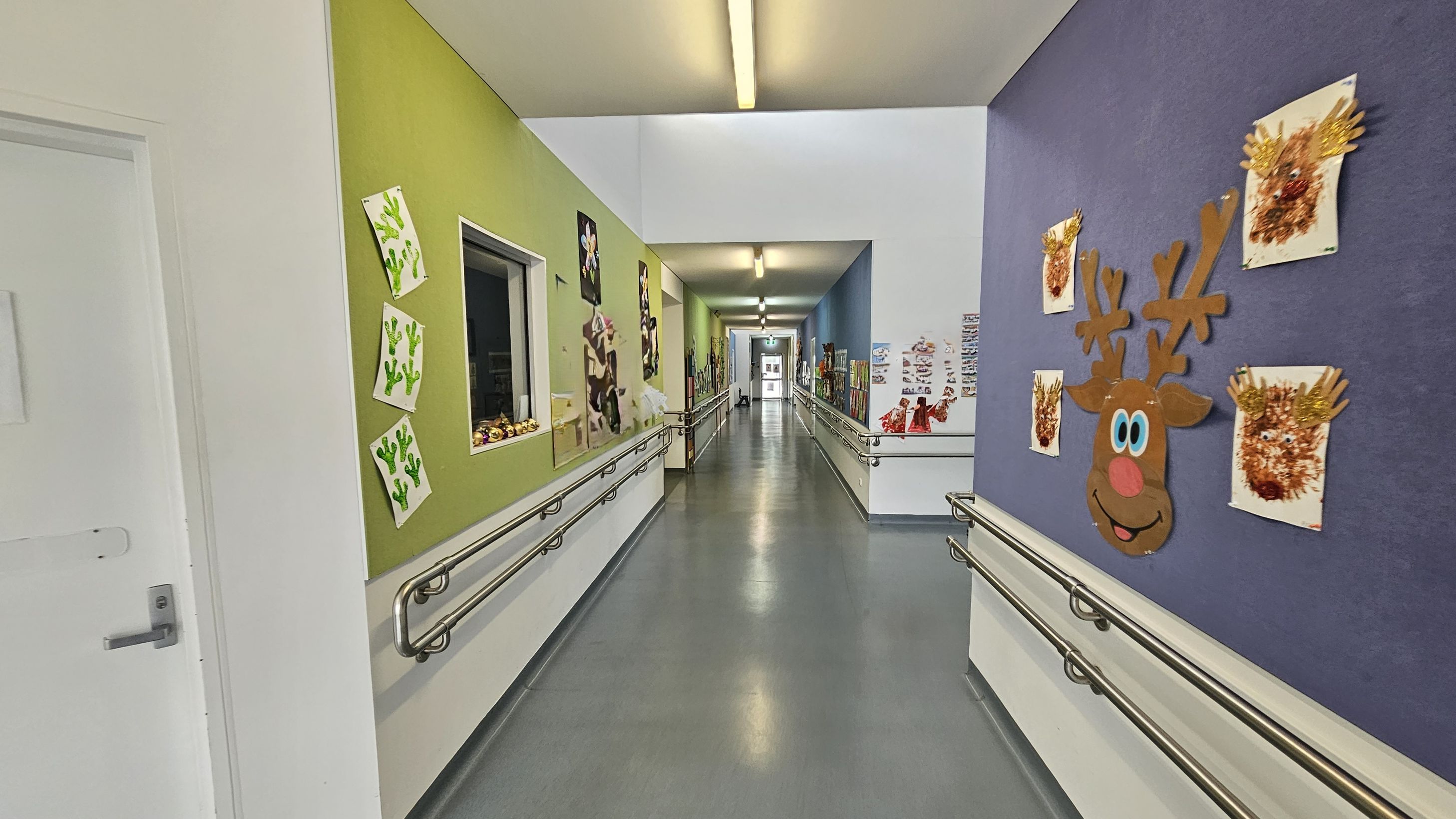 A corridor with colourful walls decorated with children's artwork. On the left, a green wall displays cactus drawings and other art pieces. On the right, a purple wall features a large reindeer face and handprint art. The floor is grey and smooth, with metal handrails on both sides. The ceiling is white with fluorescent lights. The corridor extends into the distance, with more artwork visible along the walls. A corridor with colourful walls decorated with children's artwork. On the left, a green wall displays cactus drawings and other art pieces. On the right, a purple wall features a large reindeer face and handprint art. The floor is grey and smooth, with metal handrails on both sides. The ceiling is white with fluorescent lights. The corridor extends into the distance, with more artwork visible along the walls.