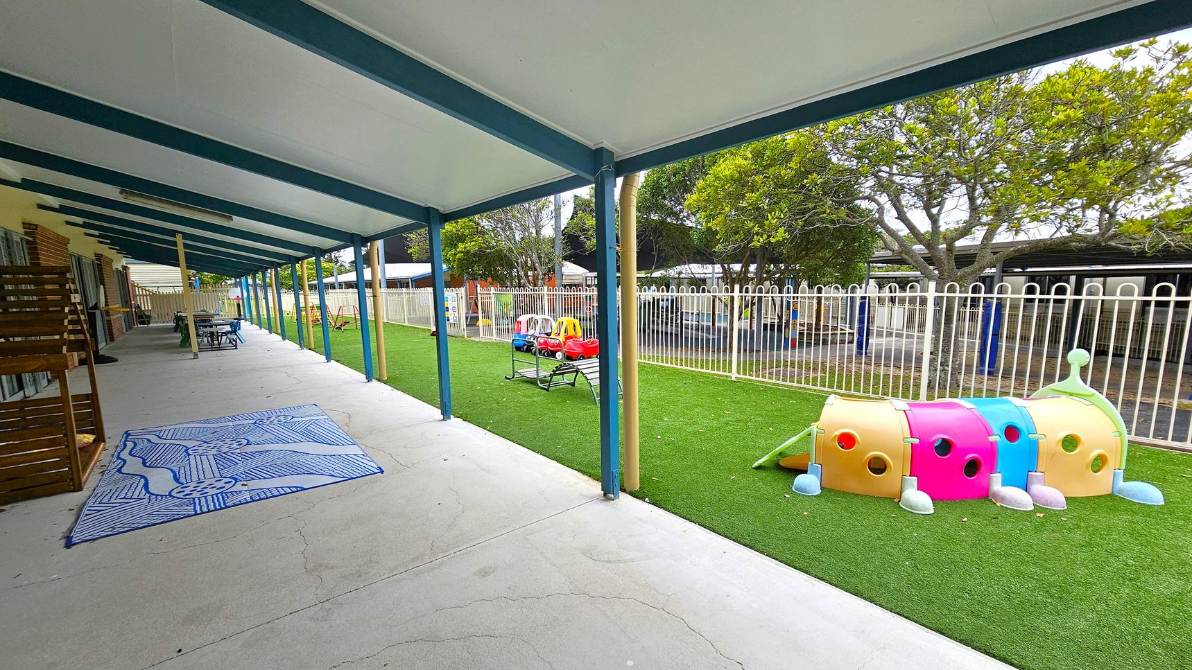 A covered outdoor area with a concrete floor and a blue and white patterned mat on the left. Wooden furniture and tables are arranged along the wall. On the right, a grassy area features colourful play equipment, including a caterpillar tunnel and toy cars. A white fence separates the play area from trees and additional structures in the background. The ceiling is supported by blue beams. A covered outdoor area with a concrete floor and a blue and white patterned mat on the left. Wooden furniture and tables are arranged along the wall. On the right, a grassy area features colourful play equipment, including a caterpillar tunnel and toy cars. A white fence separates the play area from trees and additional structures in the background. The ceiling is supported by blue beams.