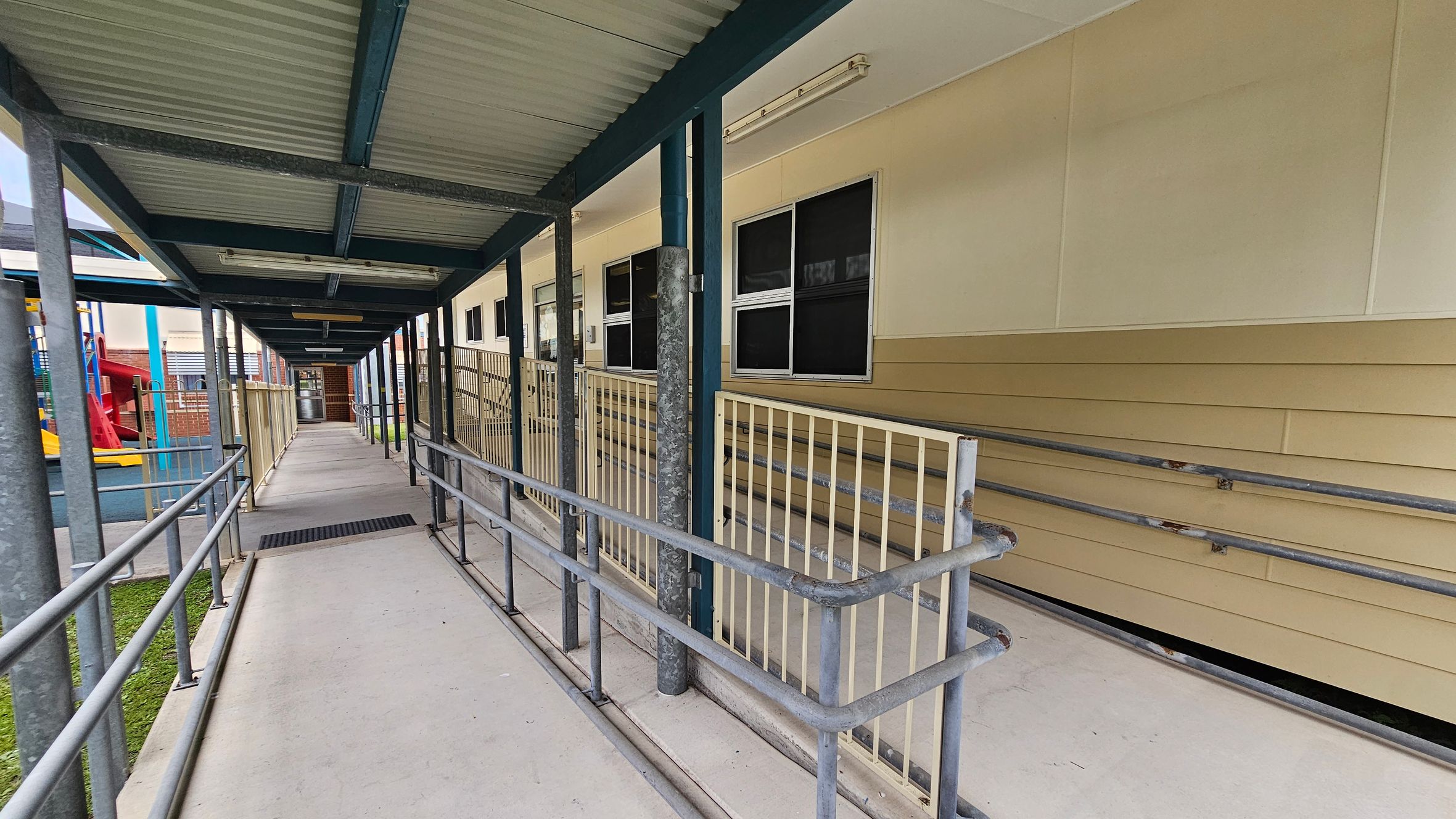 A covered walkway with a corrugated metal roof and metal railings on both sides. On the right, there is a beige building with several windows and a ramp leading up to it. On the left, a playground area is visible with a red slide and blue play structures. The floor is concrete, and the ceiling is supported by metal beams. The walkway extends into the distance, leading to a door at the far end. A covered walkway with a corrugated metal roof and metal railings on both sides. On the right, there is a beige building with several windows and a ramp leading up to it. On the left, a playground area is visible with a red slide and blue play structures. The floor is concrete, and the ceiling is supported by metal beams. The walkway extends into the distance, leading to a door at the far end.