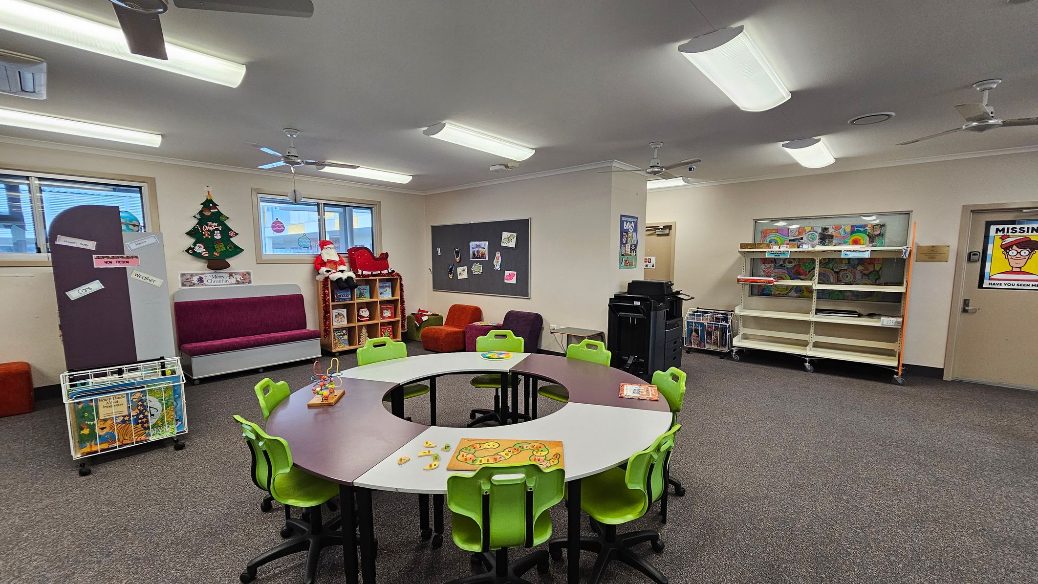 A classroom with a circular table in the centre surrounded by green chairs. On the table are educational toys and games. To the left, a purple bench sits beneath a window, with a Christmas tree decoration and a Santa figure on a shelf. The right side has a large bulletin board with artwork and a printer nearby. Shelves with books and a "Missing" poster are on the far right wall. The ceiling has fluorescent lights and fans, and the floor is carpeted. A classroom with a circular table in the centre surrounded by green chairs. On the table are educational toys and games. To the left, a purple bench sits beneath a window, with a Christmas tree decoration and a Santa figure on a shelf. The right side has a large bulletin board with artwork and a printer nearby. Shelves with books and a "Missing" poster are on the far right wall. The ceiling has fluorescent lights and fans, and the floor is carpeted.