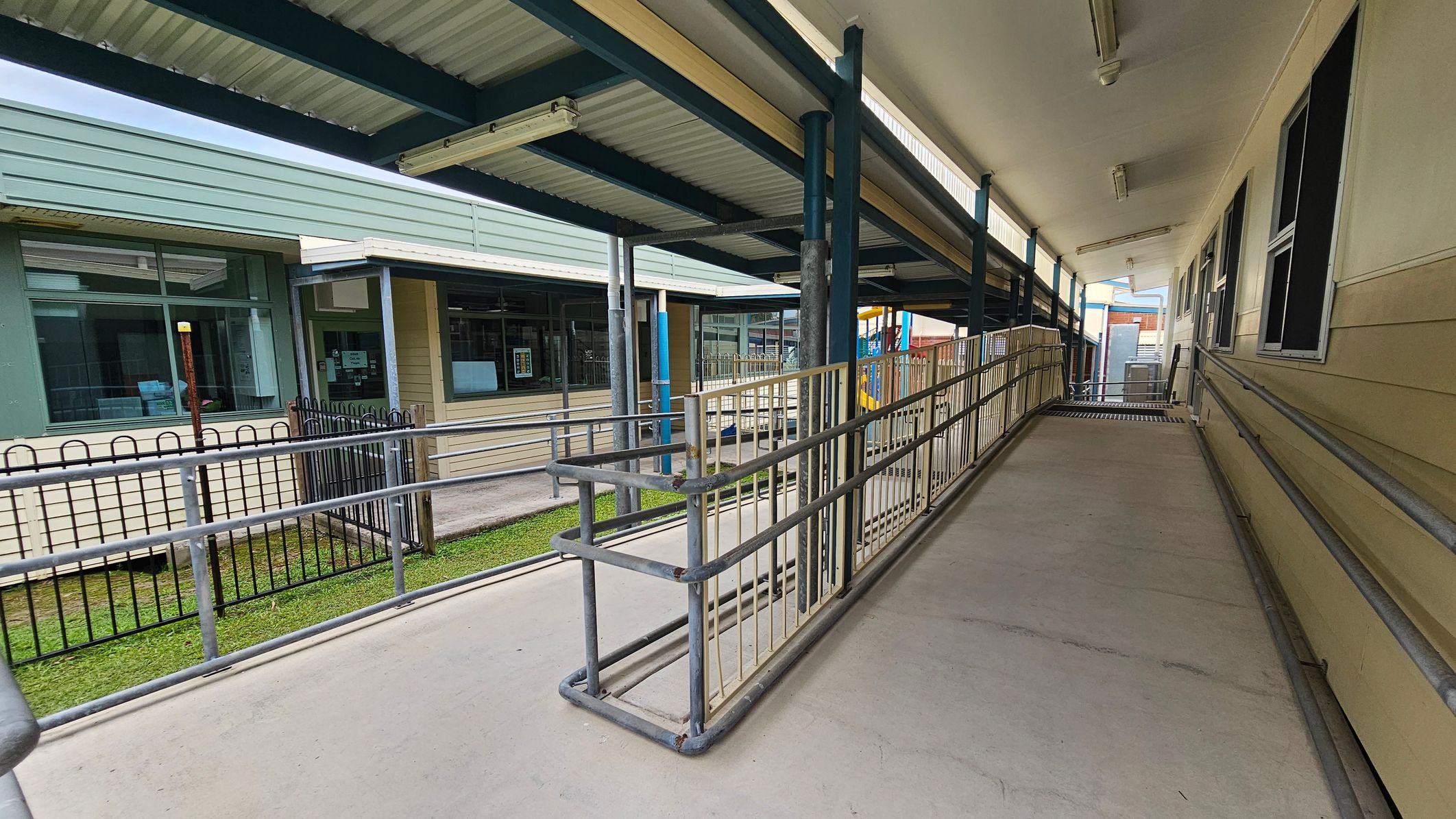 A covered walkway with a ramp and metal railings on both sides. On the right, there is a beige building with several windows. On the left, a green building with large windows and a black metal fence. The floor is concrete, and the ceiling is made of corrugated metal with fluorescent lights. In the background, there are more structures and a glimpse of a playground with colourful equipment. A covered walkway with a ramp and metal railings on both sides. On the right, there is a beige building with several windows. On the left, a green building with large windows and a black metal fence. The floor is concrete, and the ceiling is made of corrugated metal with fluorescent lights. In the background, there are more structures and a glimpse of a playground with colourful equipment.