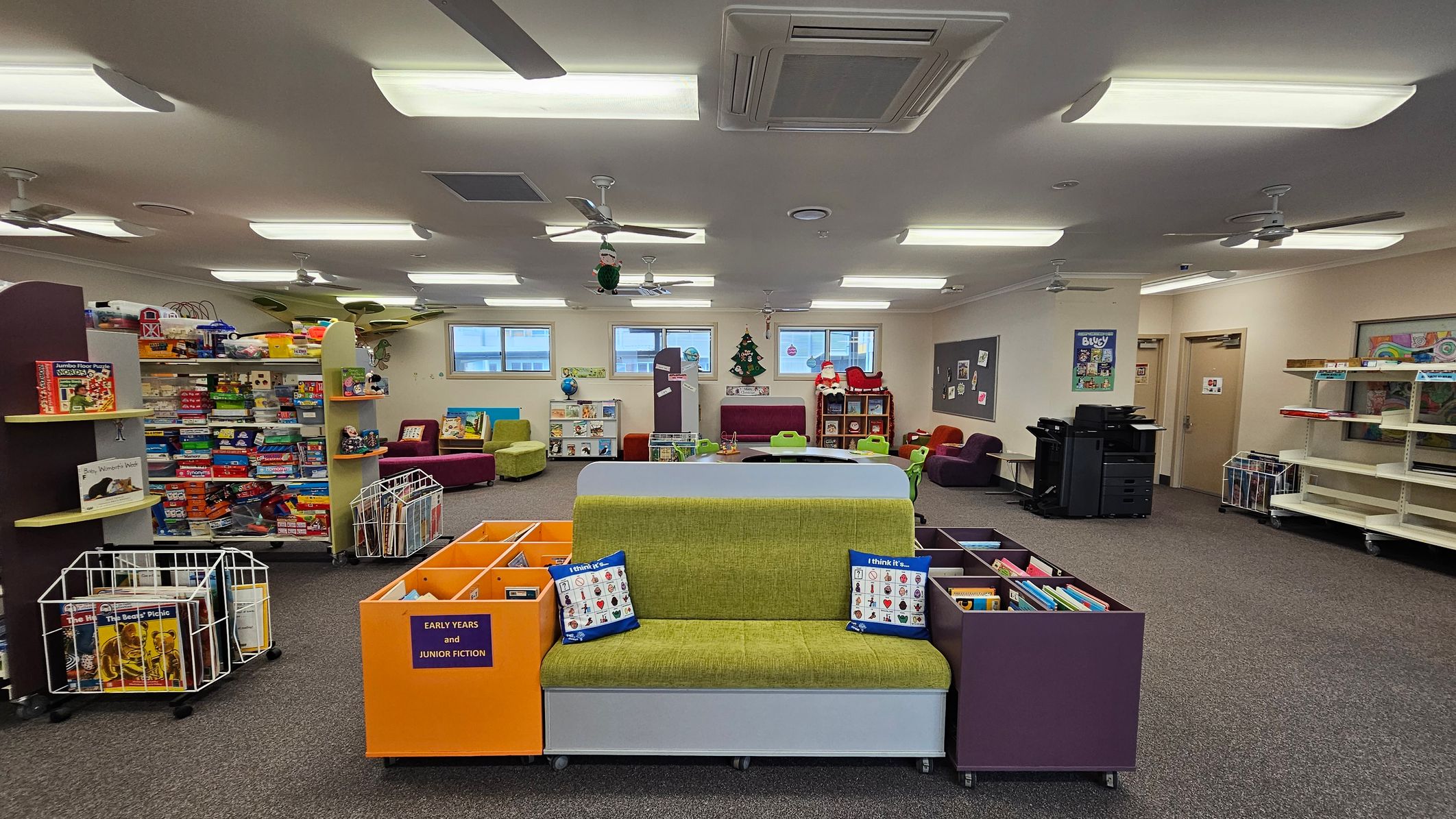 A spacious room with a carpeted floor and a ceiling fitted with fluorescent lights and ceiling fans. In the foreground, a green cushioned bench is flanked by colourful book storage units labelled "Early Years and Junior Fiction." To the left, shelves are filled with board games and educational toys. The background features windows, a Christmas tree decoration, and a Santa figure. On the right, there are printers and empty shelves. The room has a bright and organised appearance. A spacious room with a carpeted floor and a ceiling fitted with fluorescent lights and ceiling fans. In the foreground, a green cushioned bench is flanked by colourful book storage units labelled "Early Years and Junior Fiction." To the left, shelves are filled with board games and educational toys. The background features windows, a Christmas tree decoration, and a Santa figure. On the right, there are printers and empty shelves. The room has a bright and organised appearance.