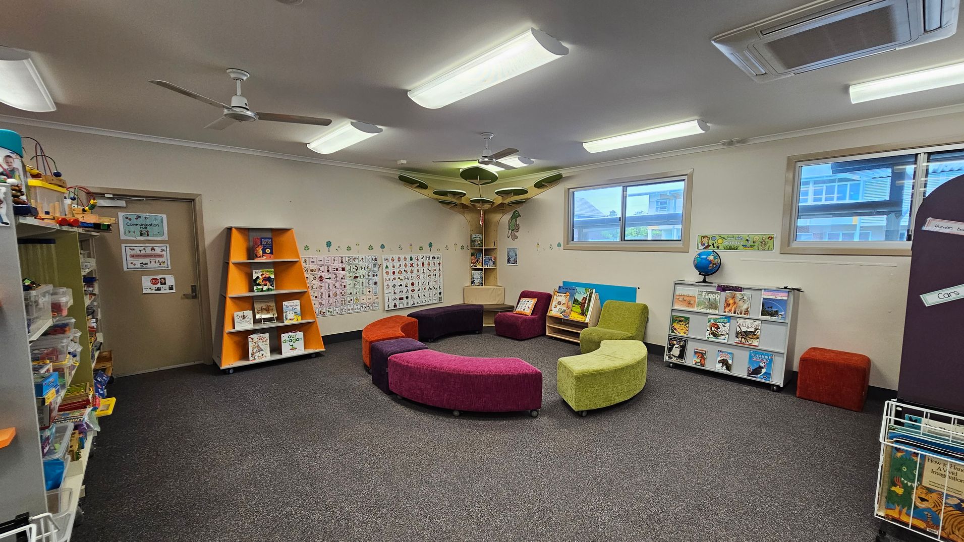 A classroom with a grey carpeted floor and a ceiling with fluorescent lights and ceiling fans. In the centre, there are colourful, curved seating pieces arranged in a circle. On the left, shelves hold various educational materials and toys. The back wall features a door and an orange triangular bookshelf with books. The right side has windows, a globe on a low shelf with books, and a small book display. The walls are decorated with educational posters and artwork. A classroom with a grey carpeted floor and a ceiling with fluorescent lights and ceiling fans. In the centre, there are colourful, curved seating pieces arranged in a circle. On the left, shelves hold various educational materials and toys. The back wall features a door and an orange triangular bookshelf with books. The right side has windows, a globe on a low shelf with books, and a small book display. The walls are decorated with educational posters and artwork.