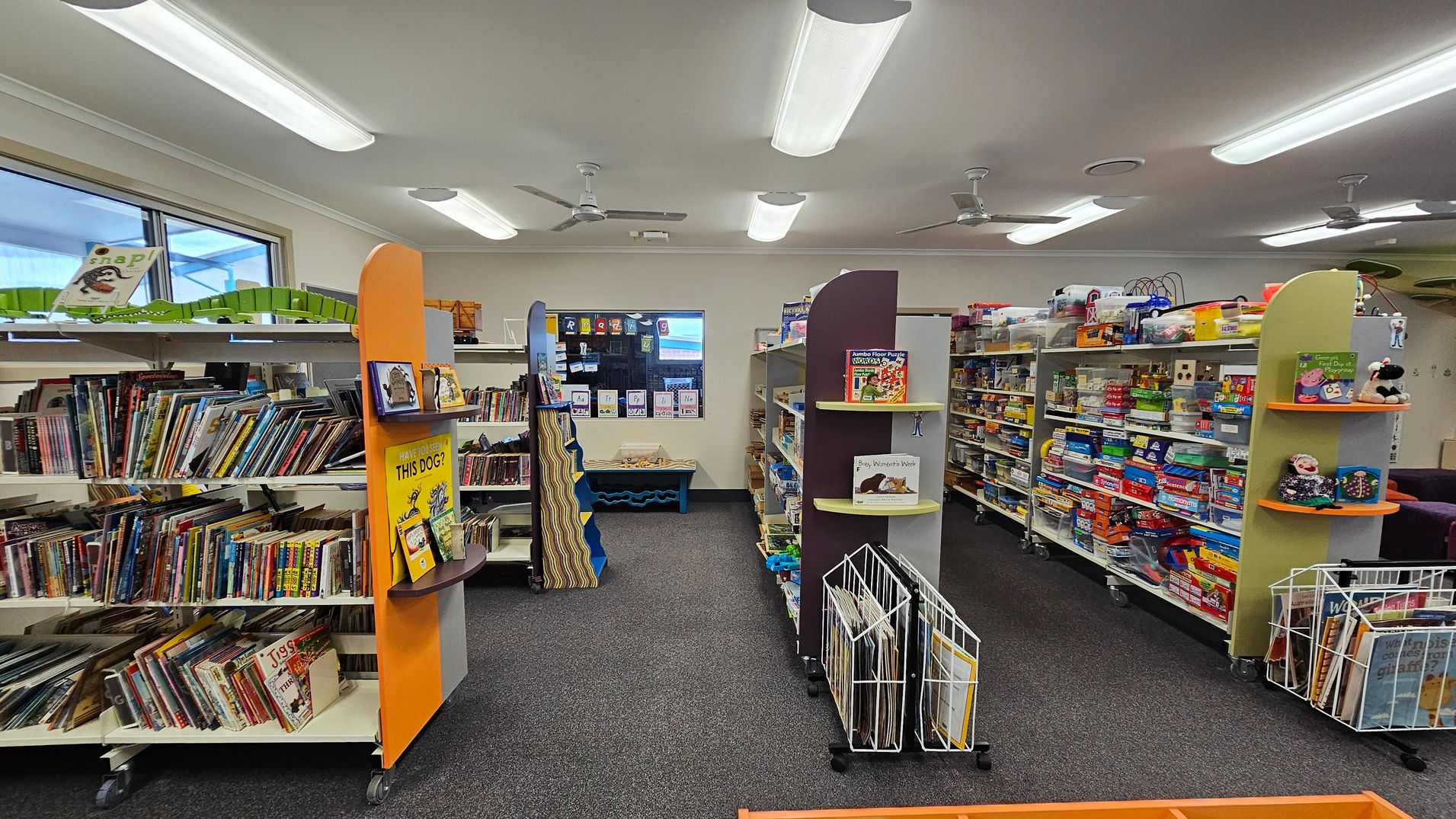 Bookshelves filled with children's books and games line both sides of a library room. On the left, shelves are packed with colourful books, while the right side features games and educational toys. In the centre, a small display shelf holds a few featured books. The floor is carpeted in grey, and the ceiling has fluorescent lights and ceiling fans. A window at the back shows a glimpse of the outside, and there are educational posters on the walls. Bookshelves filled with children's books and games line both sides of a library room. On the left, shelves are packed with colourful books, while the right side features games and educational toys. In the centre, a small display shelf holds a few featured books. The floor is carpeted in grey, and the ceiling has fluorescent lights and ceiling fans. A window at the back shows a glimpse of the outside, and there are educational posters on the walls.