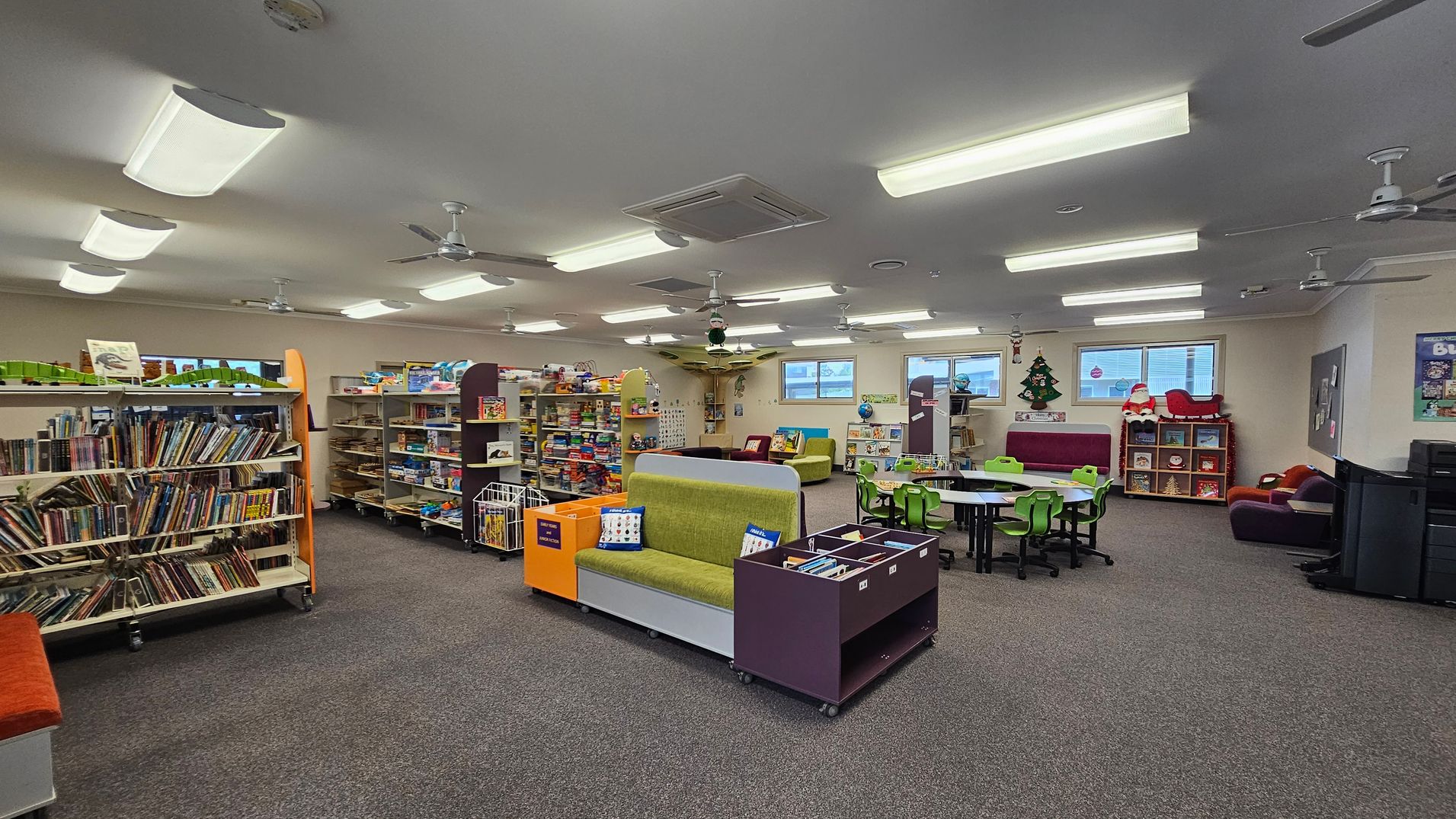 A spacious library room with grey carpeted flooring and a ceiling fitted with fluorescent lights and ceiling fans. On the left, there are bookshelves filled with books. In the centre, a green sofa with colourful cushions is flanked by storage units. To the right, there are tables with green chairs and a small seating area with plush toys. The background features windows and festive decorations, including a Christmas tree and Santa figure. A spacious library room with grey carpeted flooring and a ceiling fitted with fluorescent lights and ceiling fans. On the left, there are bookshelves filled with books. In the centre, a green sofa with colourful cushions is flanked by storage units. To the right, there are tables with green chairs and a small seating area with plush toys. The background features windows and festive decorations, including a Christmas tree and Santa figure.