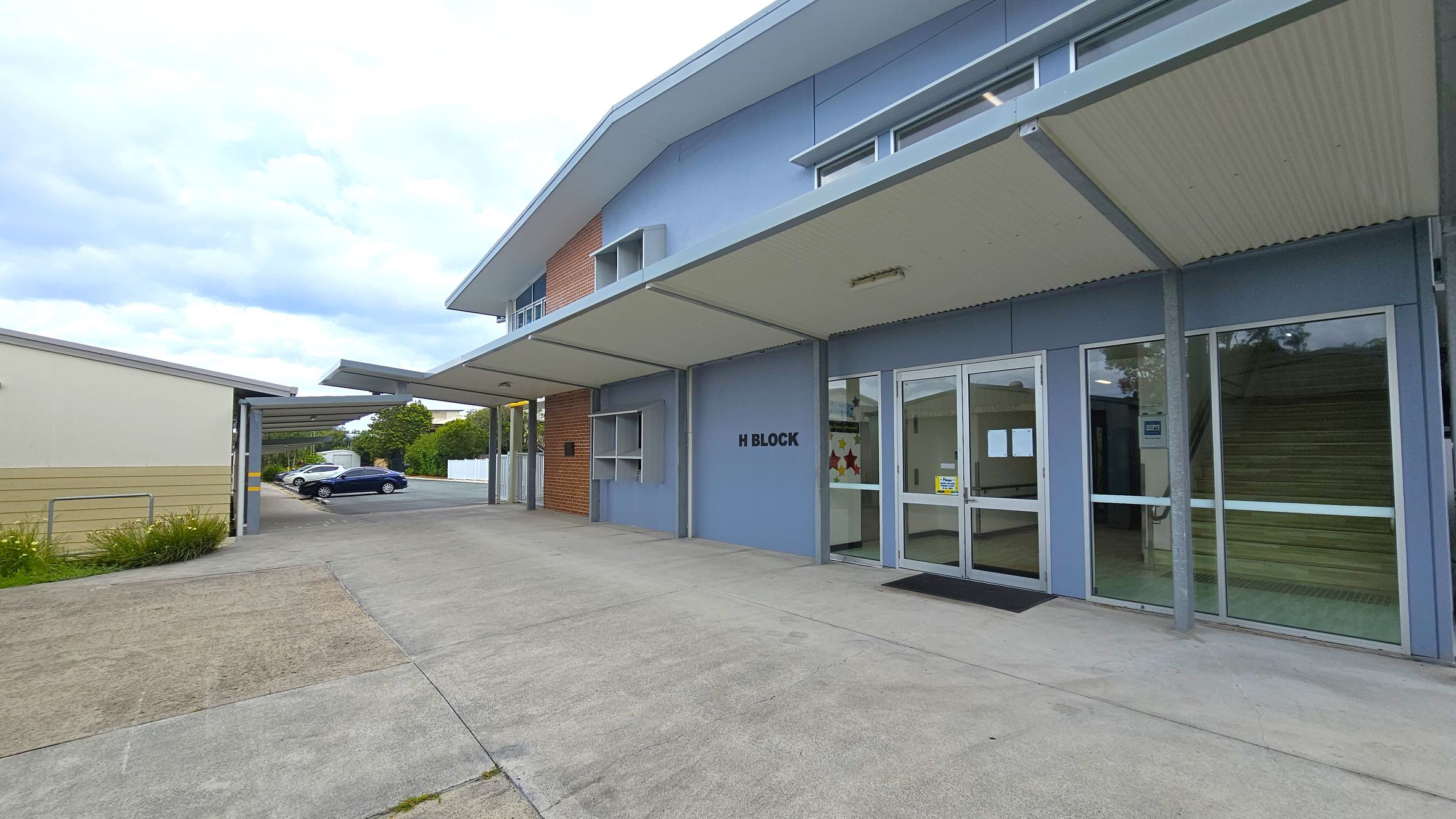 A modern building labelled "H Block" with a light blue exterior and large glass doors on the right. The building has a sloped roof with an overhang. To the left, there is a covered walkway leading to a parking area with two cars, one white and one black. The ground is paved with concrete, and there is a small patch of grass near the walkway. The background features trees and a white fence. A modern building labelled "H Block" with a light blue exterior and large glass doors on the right. The building has a sloped roof with an overhang. To the left, there is a covered walkway leading to a parking area with two cars, one white and one black. The ground is paved with concrete, and there is a small patch of grass near the walkway. The background features trees and a white fence.