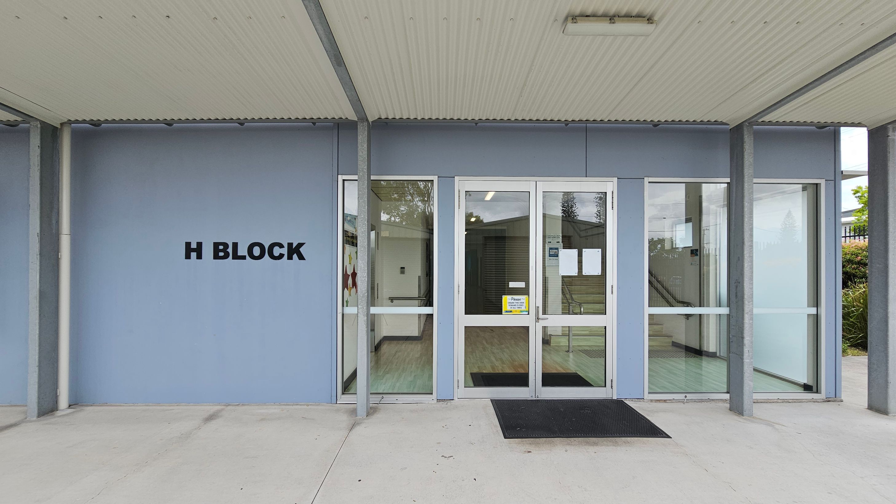 Entrance to a building labelled "H Block" on the left wall. The facade is light blue with large glass doors and windows. A black mat lies in front of the entrance. Inside, a corridor with a light-coloured floor is visible. The ceiling is corrugated and light-coloured, supported by metal beams. On the right, there are bushes outside the window. Entrance to a building labelled "H Block" on the left wall. The facade is light blue with large glass doors and windows. A black mat lies in front of the entrance. Inside, a corridor with a light-coloured floor is visible. The ceiling is corrugated and light-coloured, supported by metal beams. On the right, there are bushes outside the window.