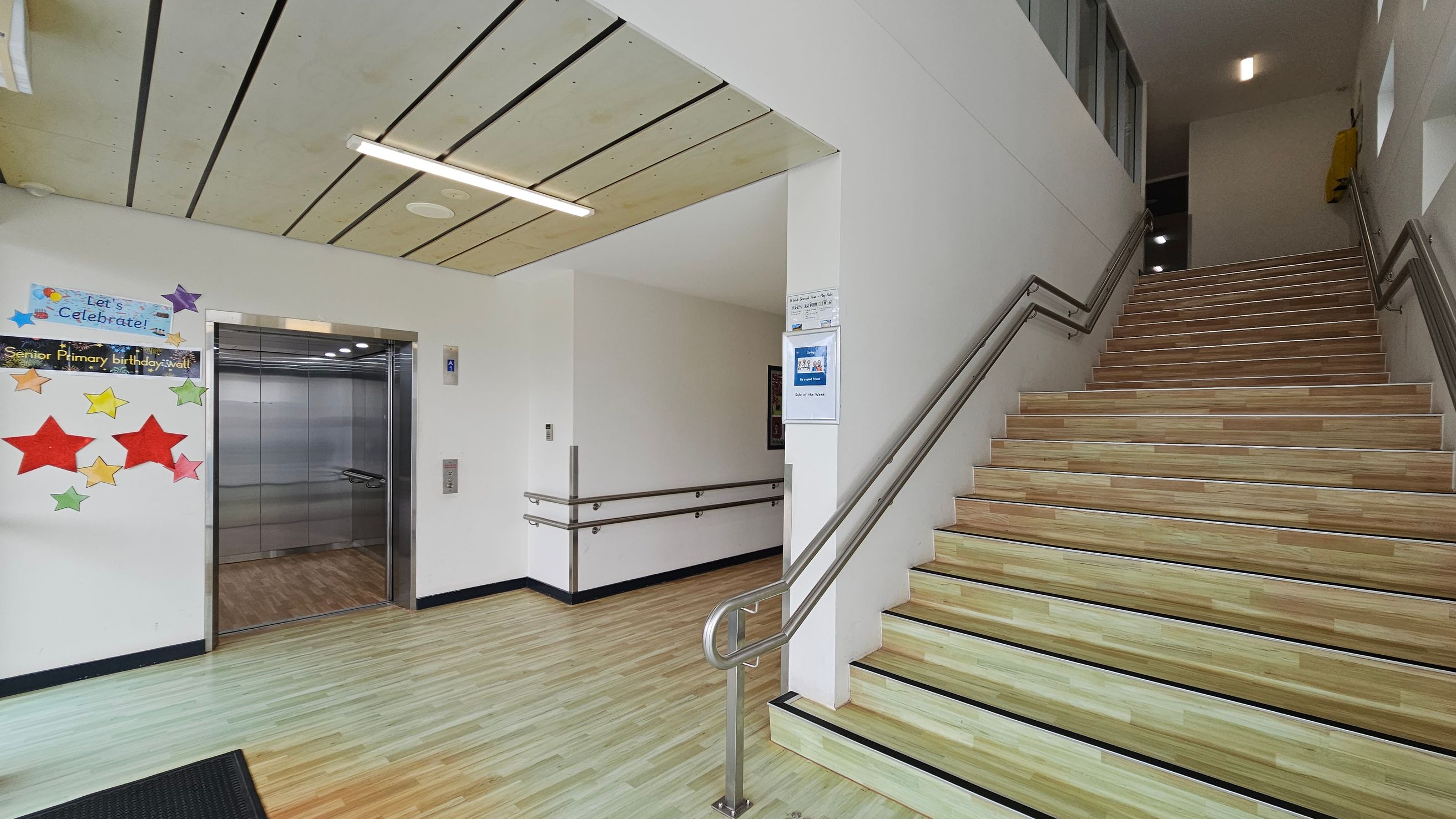 On the left, there is an open lift with a metallic interior and a colourful display of stars and a "Let's Celebrate!" sign on the wall. The floor is light wood with a black mat near the lift. On the right, a staircase with wooden steps and metal railings ascends. The ceiling is light-coloured with linear panels and a fluorescent light fixture. The walls are white, and there are informational posters near the stairs. On the left, there is an open lift with a metallic interior and a colourful display of stars and a "Let's Celebrate!" sign on the wall. The floor is light wood with a black mat near the lift. On the right, a staircase with wooden steps and metal railings ascends. The ceiling is light-coloured with linear panels and a fluorescent light fixture. The walls are white, and there are informational posters near the stairs.