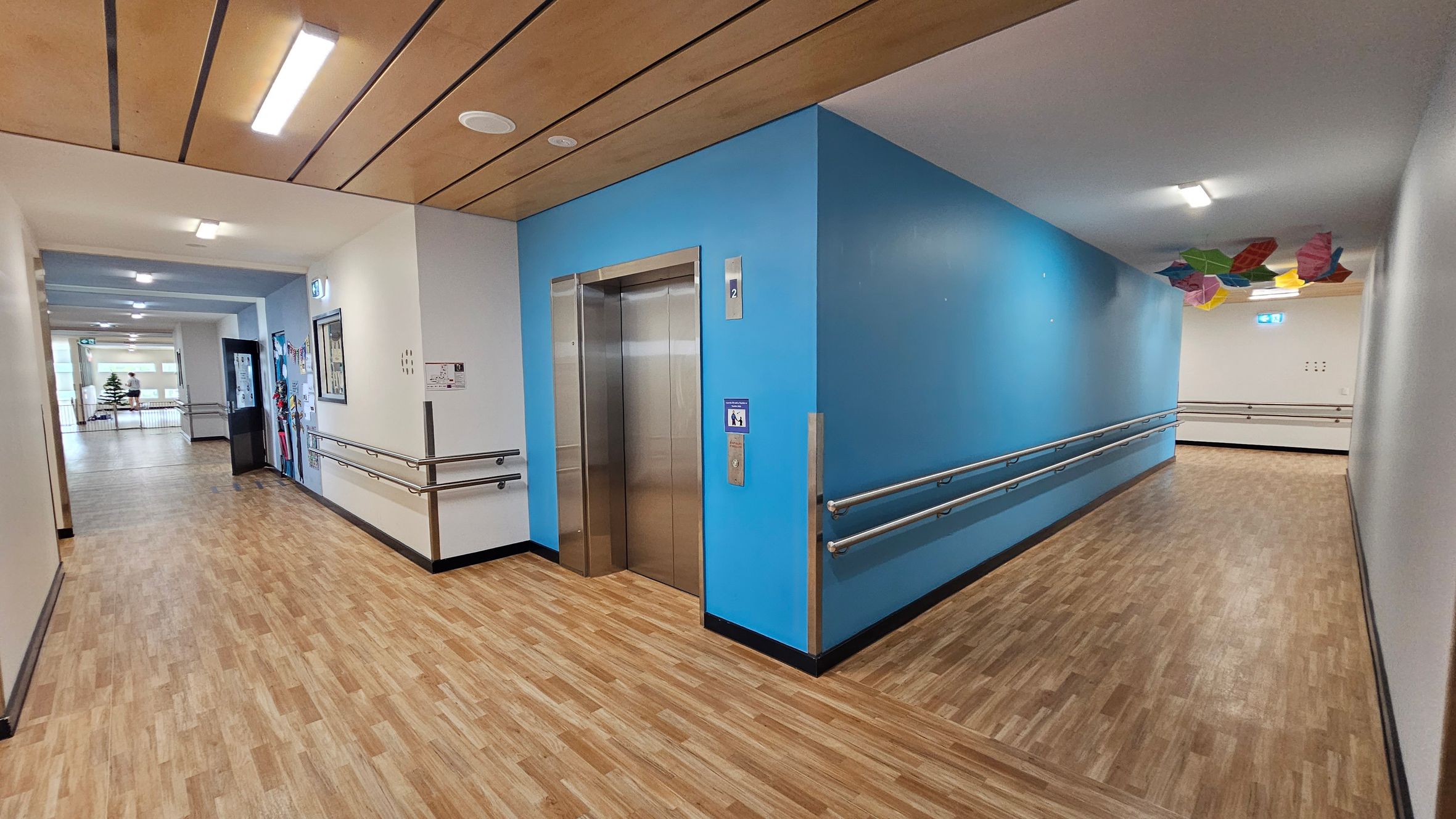 A corridor with wooden flooring and a wooden panelled ceiling. On the left, there's a lift with a blue wall and handrails. The corridor extends to the right, with colourful geometric shapes hanging from the ceiling. Walls are painted in light colours, and there are several overhead lights. A corridor with wooden flooring and a wooden panelled ceiling. On the left, there's a lift with a blue wall and handrails. The corridor extends to the right, with colourful geometric shapes hanging from the ceiling. Walls are painted in light colours, and there are several overhead lights.