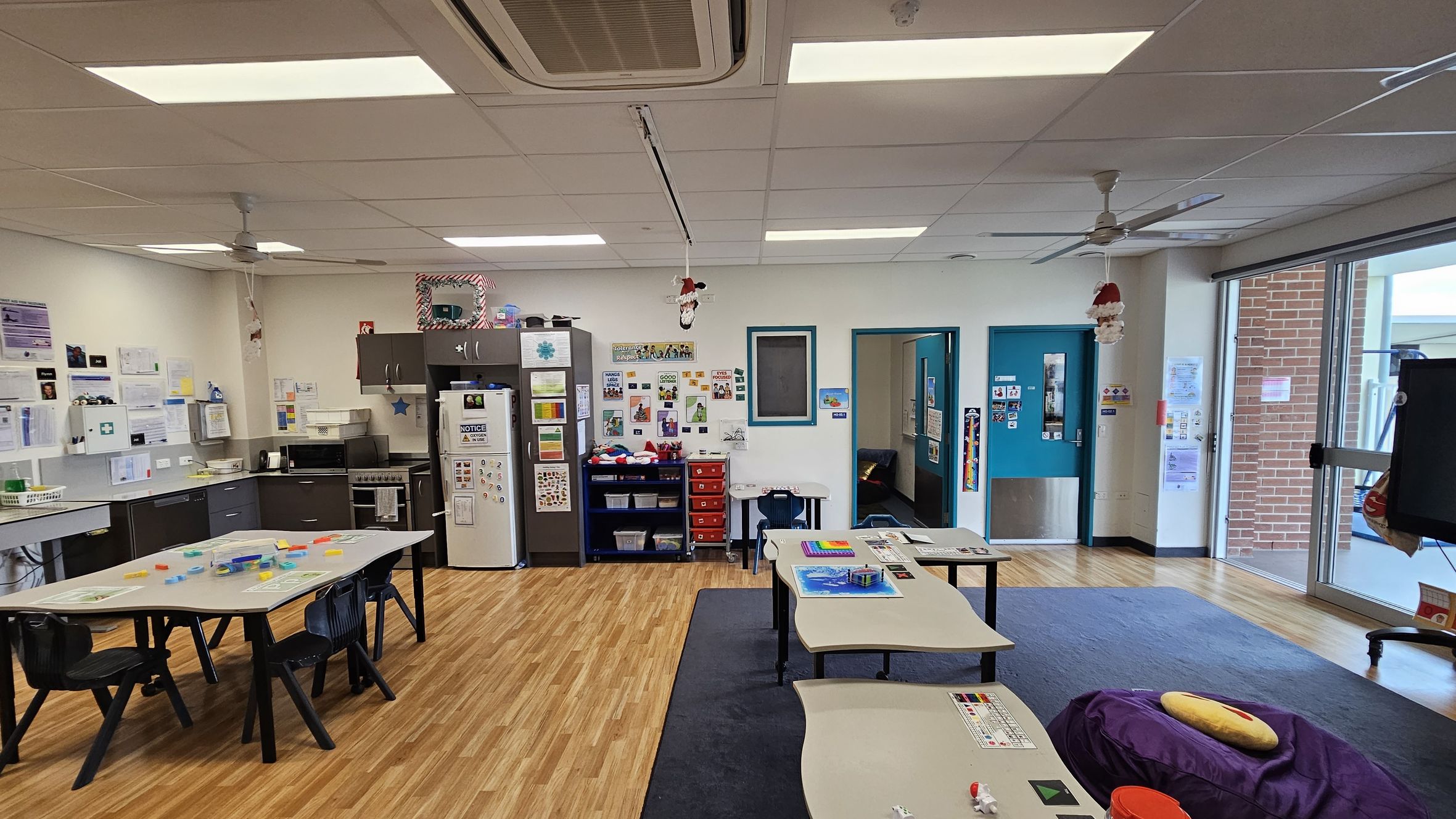 A classroom with a wooden floor and a white ceiling featuring fluorescent lights and ceiling fans. On the left, there is a kitchen area with a counter, sink, and appliances, including a fridge covered with notices. The back wall has educational posters and a small red drawer unit. In the centre, tables with chairs are arranged, some with educational materials. On the right, large windows and a door lead outside, with a brick wall visible through the glass. A classroom with a wooden floor and a white ceiling featuring fluorescent lights and ceiling fans. On the left, there is a kitchen area with a counter, sink, and appliances, including a fridge covered with notices. The back wall has educational posters and a small red drawer unit. In the centre, tables with chairs are arranged, some with educational materials. On the right, large windows and a door lead outside, with a brick wall visible through the glass.