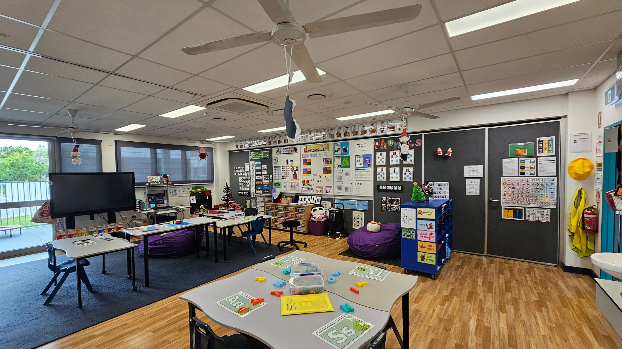 A classroom with a wooden floor and a white ceiling featuring ceiling fans. On the left, there are tables with educational materials and a large screen near windows with blinds. The middle has a wall filled with educational posters and a small Christmas tree. On the right, there are dark sliding doors with more posters, a yellow hat, and a fire extinguisher. A blue storage unit and bean bags are also visible. A classroom with a wooden floor and a white ceiling featuring ceiling fans. On the left, there are tables with educational materials and a large screen near windows with blinds. The middle has a wall filled with educational posters and a small Christmas tree. On the right, there are dark sliding doors with more posters, a yellow hat, and a fire extinguisher. A blue storage unit and bean bags are also visible.