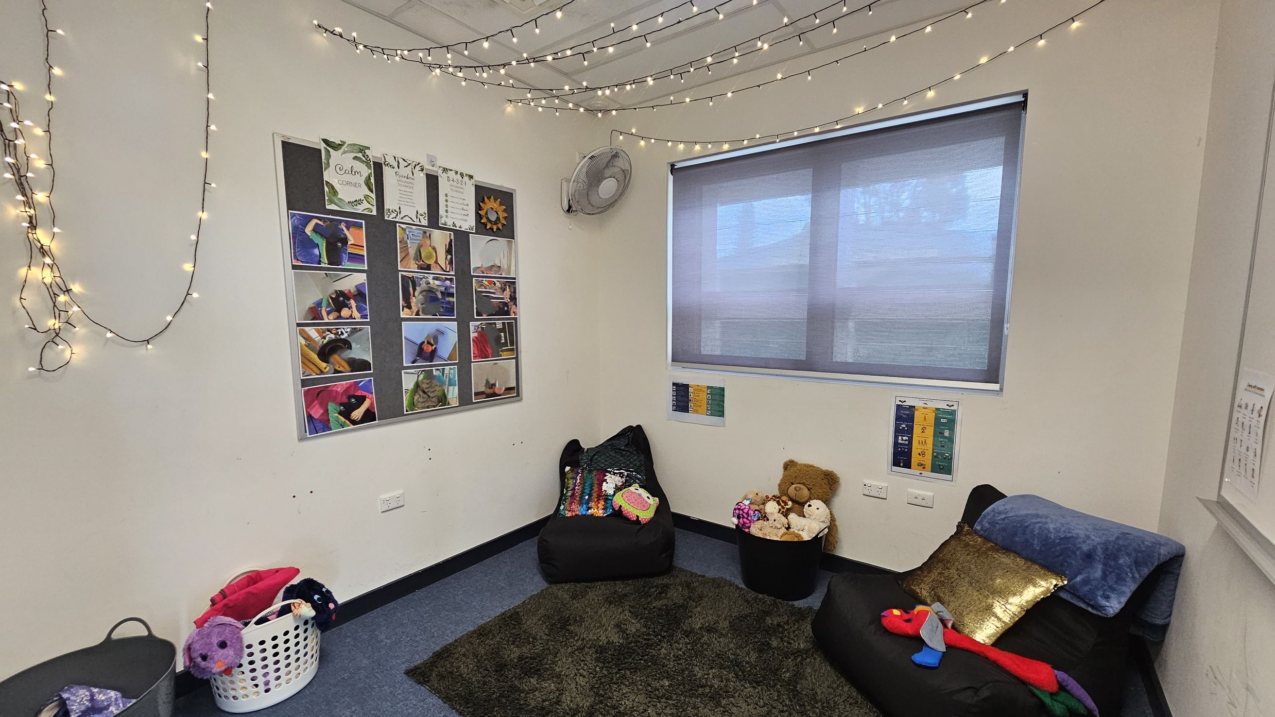 A cosy room with a soft, dark carpet on the floor. On the left, a bulletin board displays various photos and papers. Fairy lights are strung across the ceiling. In the corner, a fan is mounted on the wall. The right side features a window with a grey blind, beneath which are two black chairs with colourful cushions and a basket of stuffed toys. A small chart is visible on the wall near the window. A laundry basket with plush toys is on the floor. A cosy room with a soft, dark carpet on the floor. On the left, a bulletin board displays various photos and papers. Fairy lights are strung across the ceiling. In the corner, a fan is mounted on the wall. The right side features a window with a grey blind, beneath which are two black chairs with colourful cushions and a basket of stuffed toys. A small chart is visible on the wall near the window. A laundry basket with plush toys is on the floor.