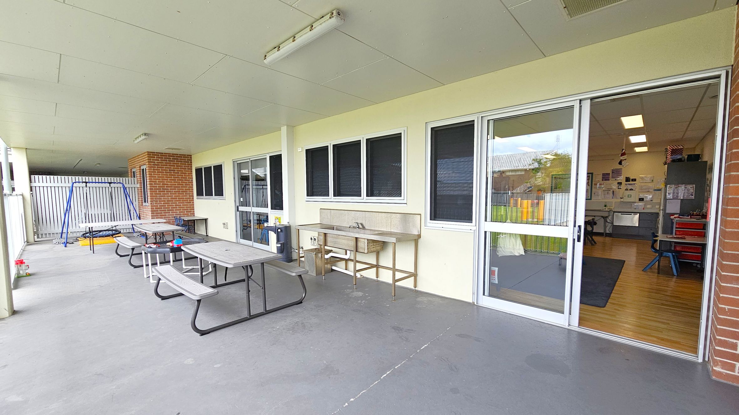 A covered outdoor area with a concrete floor and a ceiling with fluorescent lights. On the left, there is a blue swing set and several picnic tables with benches. In the middle, a stainless steel sink is mounted against the wall. On the right, large sliding glass doors lead into an indoor room with wooden flooring, visible tables, chairs, and a cabinet. The walls are a light colour, and there is a brick column near the entrance. A covered outdoor area with a concrete floor and a ceiling with fluorescent lights. On the left, there is a blue swing set and several picnic tables with benches. In the middle, a stainless steel sink is mounted against the wall. On the right, large sliding glass doors lead into an indoor room with wooden flooring, visible tables, chairs, and a cabinet. The walls are a light colour, and there is a brick column near the entrance.