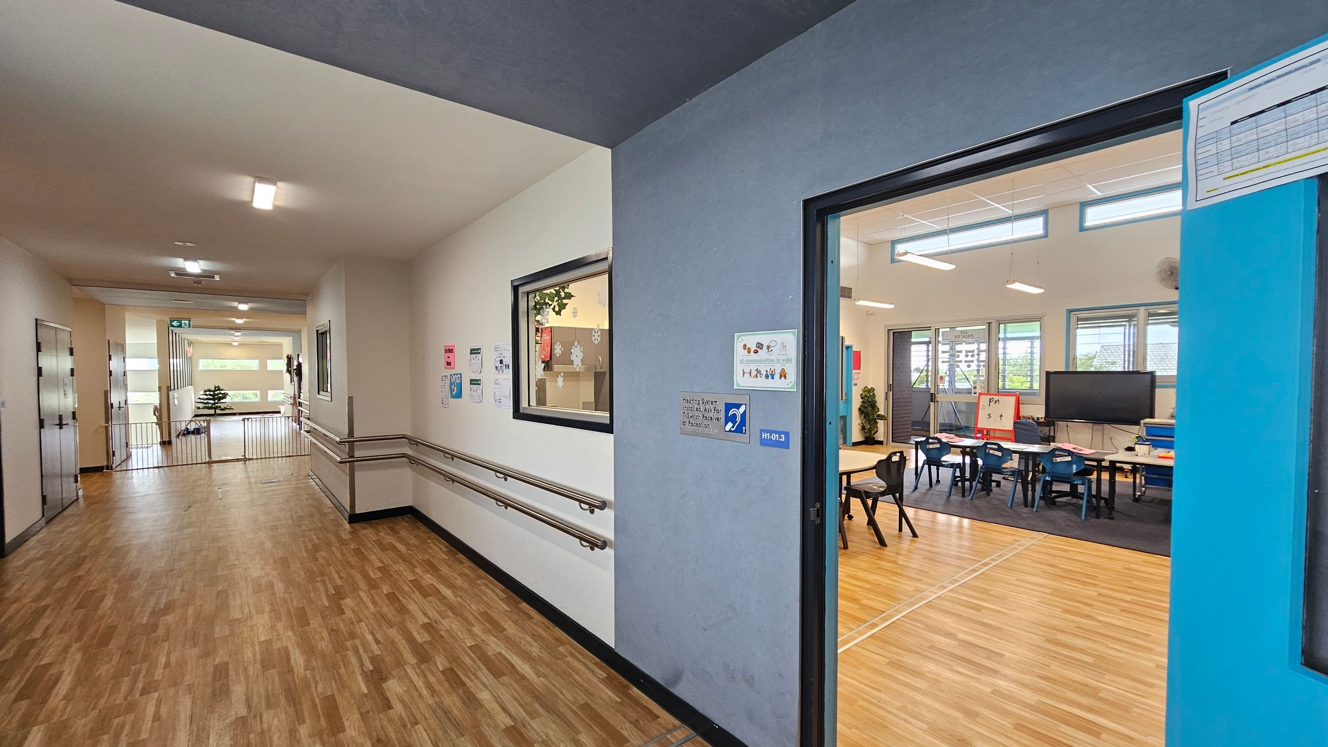 A school corridor with wooden flooring and white walls. On the left, there are handrails along the wall and a window with a view into a room. The corridor leads to a bright area with large windows and a small plant. On the right, an open door reveals a classroom with tables, chairs, a large screen, and a whiteboard. The classroom has light wooden flooring and several windows allowing natural light. Ceiling lights are visible in both the corridor and the A school corridor with wooden flooring and white walls. On the left, there are handrails along the wall and a window with a view into a room. The corridor leads to a bright area with large windows and a small plant. On the right, an open door reveals a classroom with tables, chairs, a large screen, and a whiteboard. The classroom has light wooden flooring and several windows allowing natural light. Ceiling lights are visible in both the corridor and the
