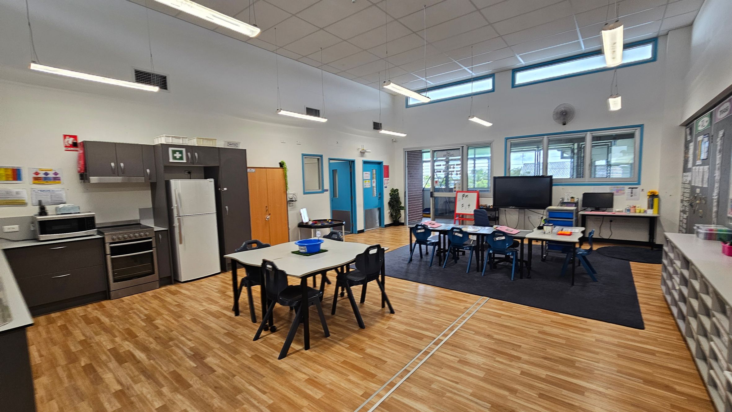 A classroom with a wooden floor and a white ceiling featuring fluorescent lights. On the left, there is a kitchen area with a stove, microwave, and refrigerator. In the centre, tables and chairs are arranged for students. The right side has a large screen, a computer on a desk, and a whiteboard. Windows line the upper part of the walls, allowing natural light. Storage units with multiple compartments are visible on the far right. A classroom with a wooden floor and a white ceiling featuring fluorescent lights. On the left, there is a kitchen area with a stove, microwave, and refrigerator. In the centre, tables and chairs are arranged for students. The right side has a large screen, a computer on a desk, and a whiteboard. Windows line the upper part of the walls, allowing natural light. Storage units with multiple compartments are visible on the far right.