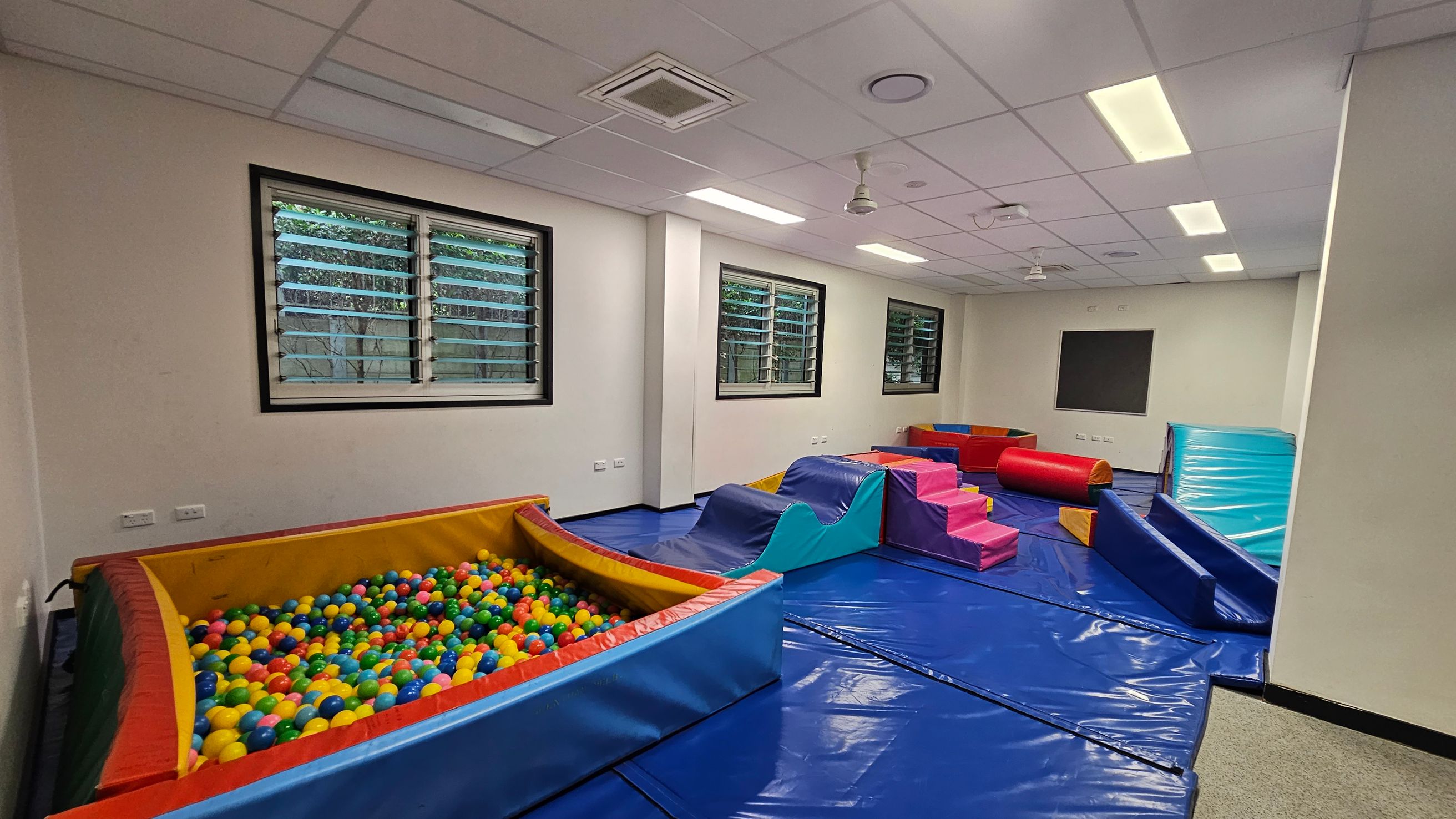 A playroom with a ball pit filled with colourful balls on the left. In the centre, there are soft play structures including a blue wave-shaped ramp and pink steps. On the right, a red cylindrical cushion and a blue mat are visible. The floor is covered with blue mats, and the ceiling has white panels with fluorescent lights. Three windows with horizontal blinds are on the back wall, allowing natural light to enter. A playroom with a ball pit filled with colourful balls on the left. In the centre, there are soft play structures including a blue wave-shaped ramp and pink steps. On the right, a red cylindrical cushion and a blue mat are visible. The floor is covered with blue mats, and the ceiling has white panels with fluorescent lights. Three windows with horizontal blinds are on the back wall, allowing natural light to enter.