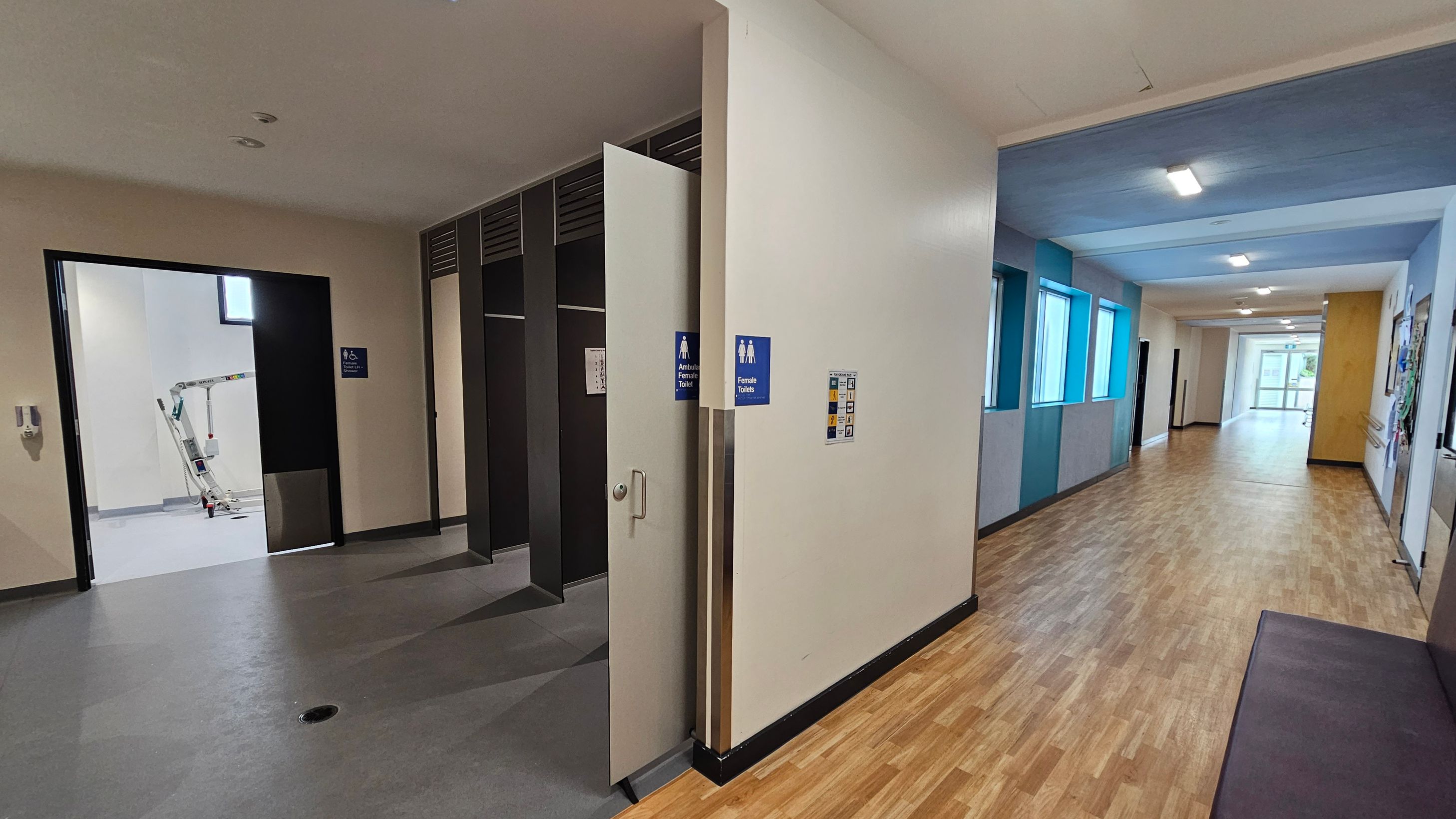 On the left, there is an open door leading to a room with a grey floor and a piece of equipment visible inside. The door has signs indicating accessible and female toilets. On the right, a corridor with a wooden floor extends into the distance, featuring several windows with blue frames on the left wall. The ceiling is white with recessed lighting. The corridor leads to a glass door at the far end. On the left, there is an open door leading to a room with a grey floor and a piece of equipment visible inside. The door has signs indicating accessible and female toilets. On the right, a corridor with a wooden floor extends into the distance, featuring several windows with blue frames on the left wall. The ceiling is white with recessed lighting. The corridor leads to a glass door at the far end.