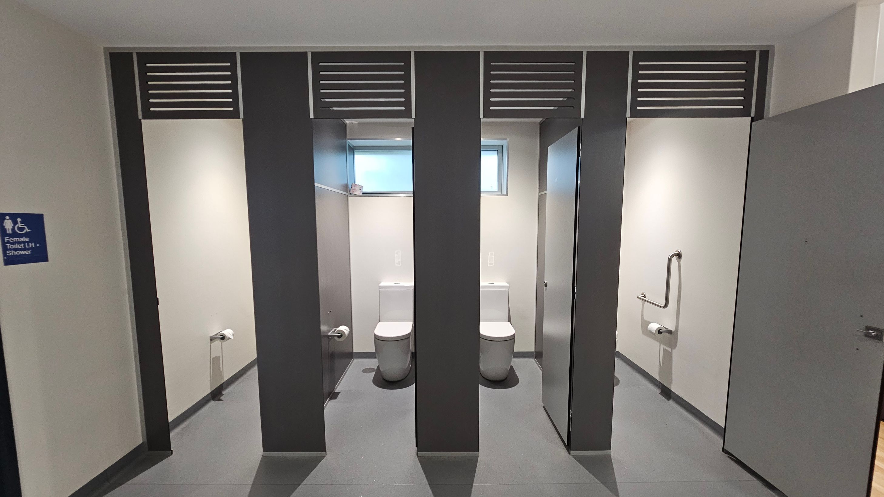 A public restroom with two toilet cubicles in the centre, each with a white toilet and a small window above. The cubicles have grey partitions with ventilation panels at the top. On the right, a door is partially open, revealing a grab bar and toilet paper holder. On the left, a sign indicates a female toilet with shower facilities. The floor is grey, and the walls are light-coloured. A public restroom with two toilet cubicles in the centre, each with a white toilet and a small window above. The cubicles have grey partitions with ventilation panels at the top. On the right, a door is partially open, revealing a grab bar and toilet paper holder. On the left, a sign indicates a female toilet with shower facilities. The floor is grey, and the walls are light-coloured.