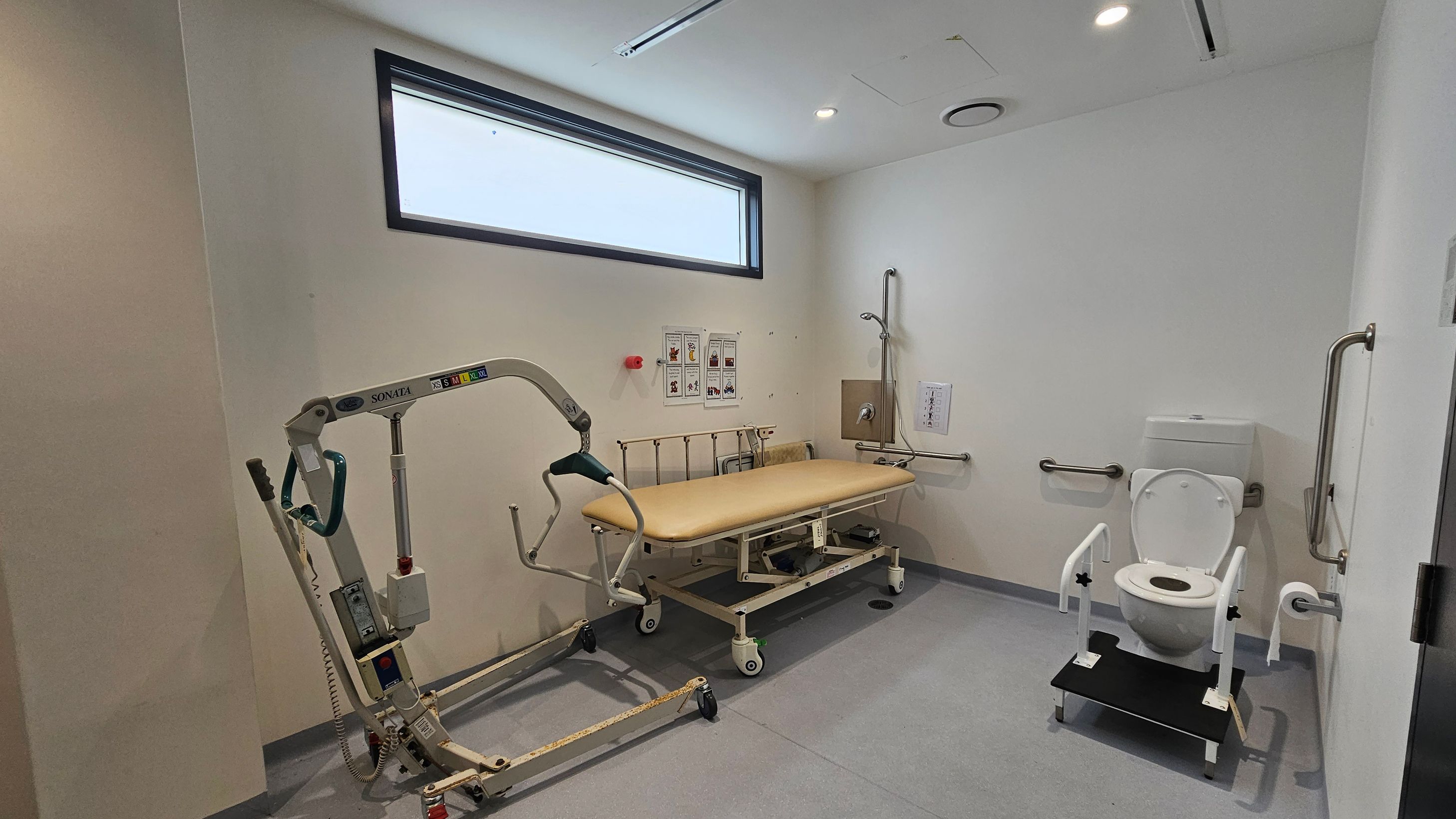 A room with a grey floor and white walls. On the left, there is a mobile hoist with a green handle. In the centre, a beige adjustable bed with side rails is positioned against the wall, beneath a rectangular window. On the right, a toilet with support bars and a foot platform is installed. The walls have several grab bars and a shower head. The ceiling is white with recessed lighting and ventilation. A room with a grey floor and white walls. On the left, there is a mobile hoist with a green handle. In the centre, a beige adjustable bed with side rails is positioned against the wall, beneath a rectangular window. On the right, a toilet with support bars and a foot platform is installed. The walls have several grab bars and a shower head. The ceiling is white with recessed lighting and ventilation.