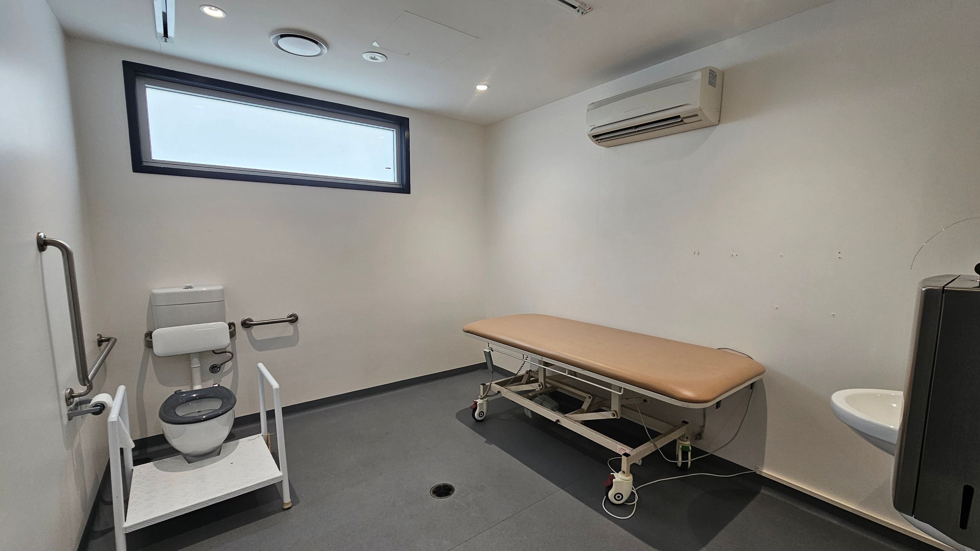 A room with a grey floor and white walls. On the left, there's a toilet with support rails and a white frame. In the middle, a brown cushioned examination table on wheels. On the right, a small white sink with a black hand dryer above it. A rectangular window is high on the left wall, and an air conditioning unit is mounted on the right wall. The ceiling has recessed lighting and ventilation fixtures. A room with a grey floor and white walls. On the left, there's a toilet with support rails and a white frame. In the middle, a brown cushioned examination table on wheels. On the right, a small white sink with a black hand dryer above it. A rectangular window is high on the left wall, and an air conditioning unit is mounted on the right wall. The ceiling has recessed lighting and ventilation fixtures.