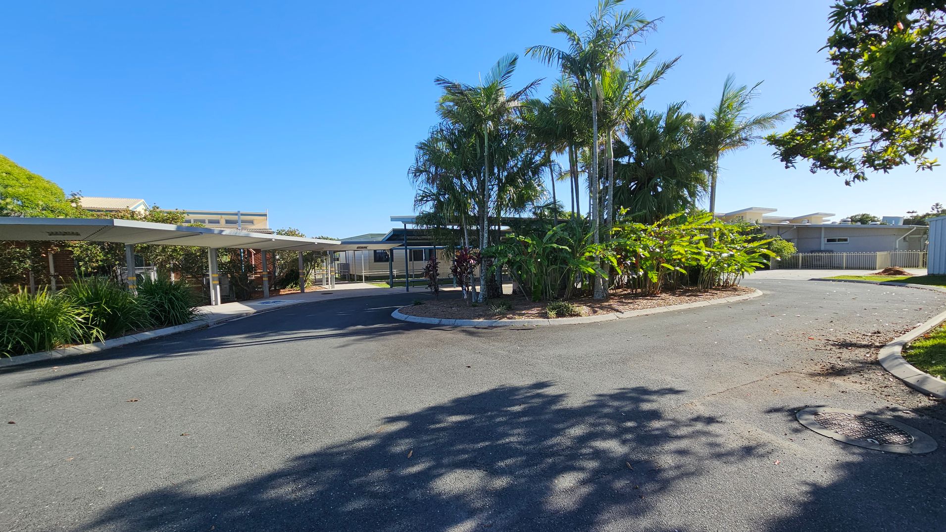 A paved driveway curves around a central island filled with tall palm trees and lush green plants. On the left, there are covered parking spaces with metal roofs, bordered by more greenery. In the background, a low building with a flat roof is visible, partially obscured by the trees. The area is well-maintained, with shadows of trees cast on the pavement. A paved driveway curves around a central island filled with tall palm trees and lush green plants. On the left, there are covered parking spaces with metal roofs, bordered by more greenery. In the background, a low building with a flat roof is visible, partially obscured by the trees. The area is well-maintained, with shadows of trees cast on the pavement.