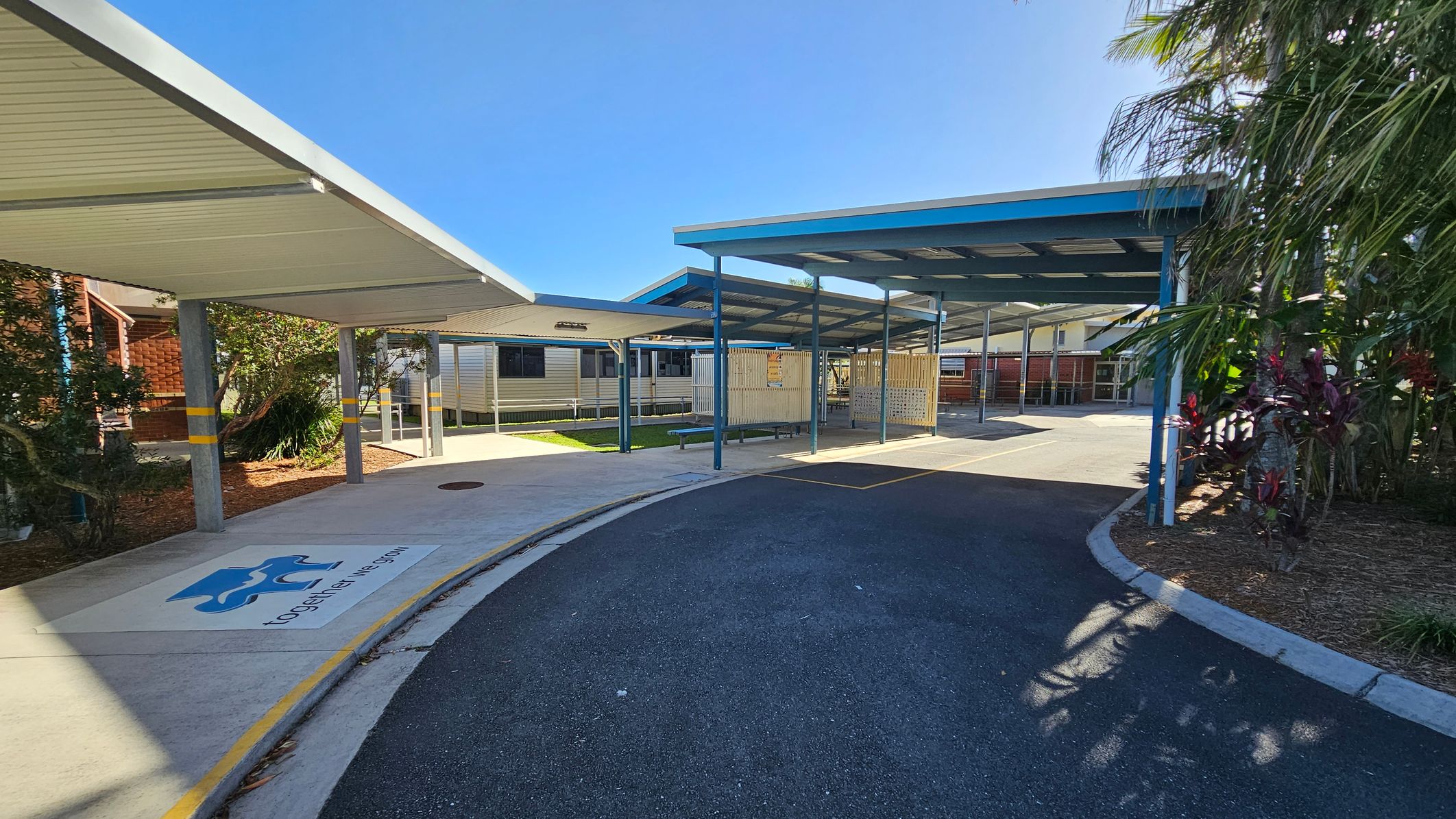 Covered walkways with blue and white roofs extend from the left to the centre, supported by grey pillars. A pathway curves from the foreground to the centre, with a logo and text on the ground. On the right, there are plants and trees. In the background, there are buildings with metal siding and windows. The ground is a mix of concrete and asphalt. Covered walkways with blue and white roofs extend from the left to the centre, supported by grey pillars. A pathway curves from the foreground to the centre, with a logo and text on the ground. On the right, there are plants and trees. In the background, there are buildings with metal siding and windows. The ground is a mix of concrete and asphalt.