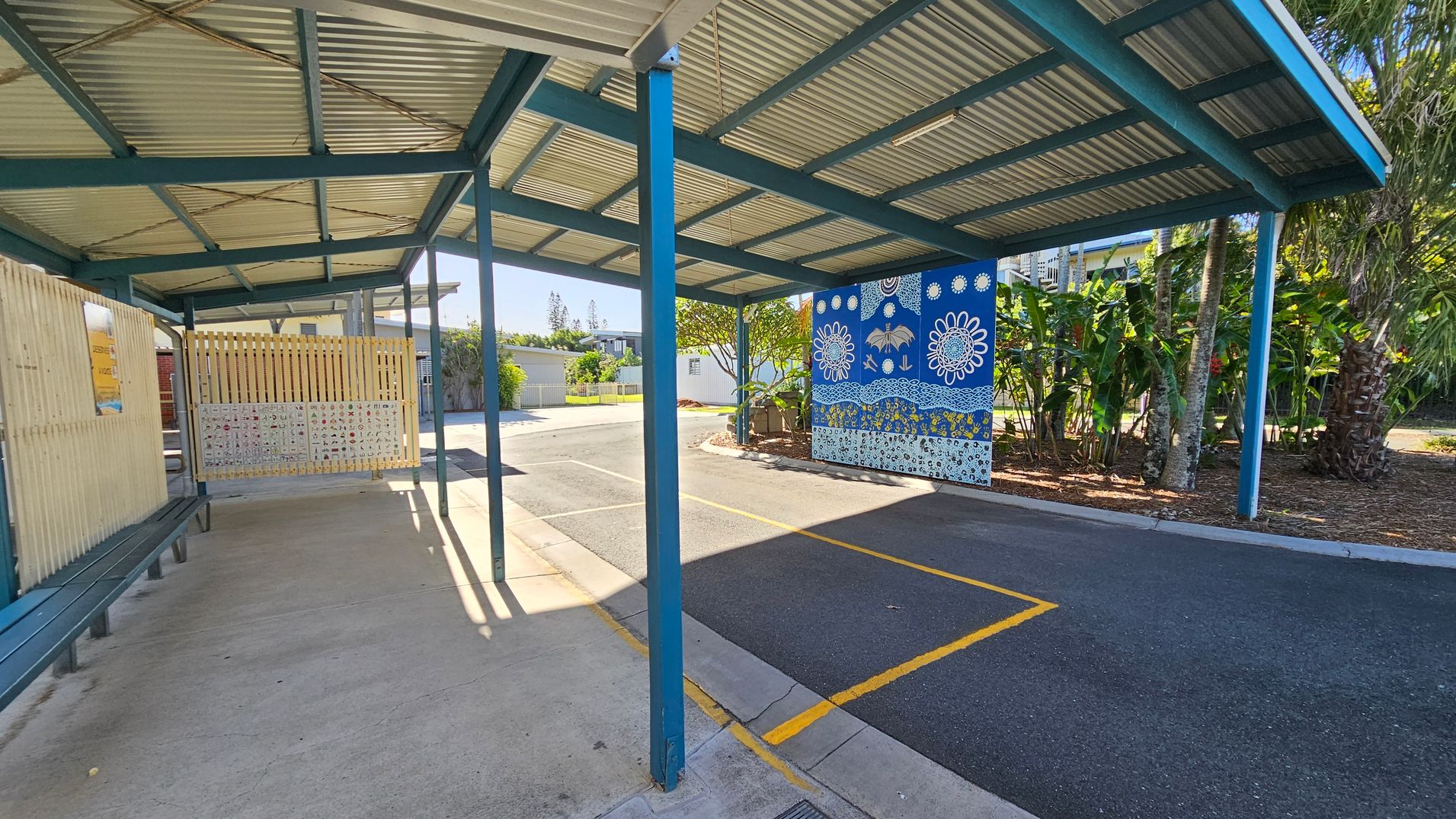 A covered walkway with a corrugated metal roof supported by blue beams. On the left, there is a wooden fence with a bench beneath it. The ground is a mix of concrete and asphalt. On the right, a vibrant blue mural with white floral and abstract designs is visible. Behind the mural, there are lush green plants and trees. The area is well-lit with sunlight casting shadows on the ground. A covered walkway with a corrugated metal roof supported by blue beams. On the left, there is a wooden fence with a bench beneath it. The ground is a mix of concrete and asphalt. On the right, a vibrant blue mural with white floral and abstract designs is visible. Behind the mural, there are lush green plants and trees. The area is well-lit with sunlight casting shadows on the ground.